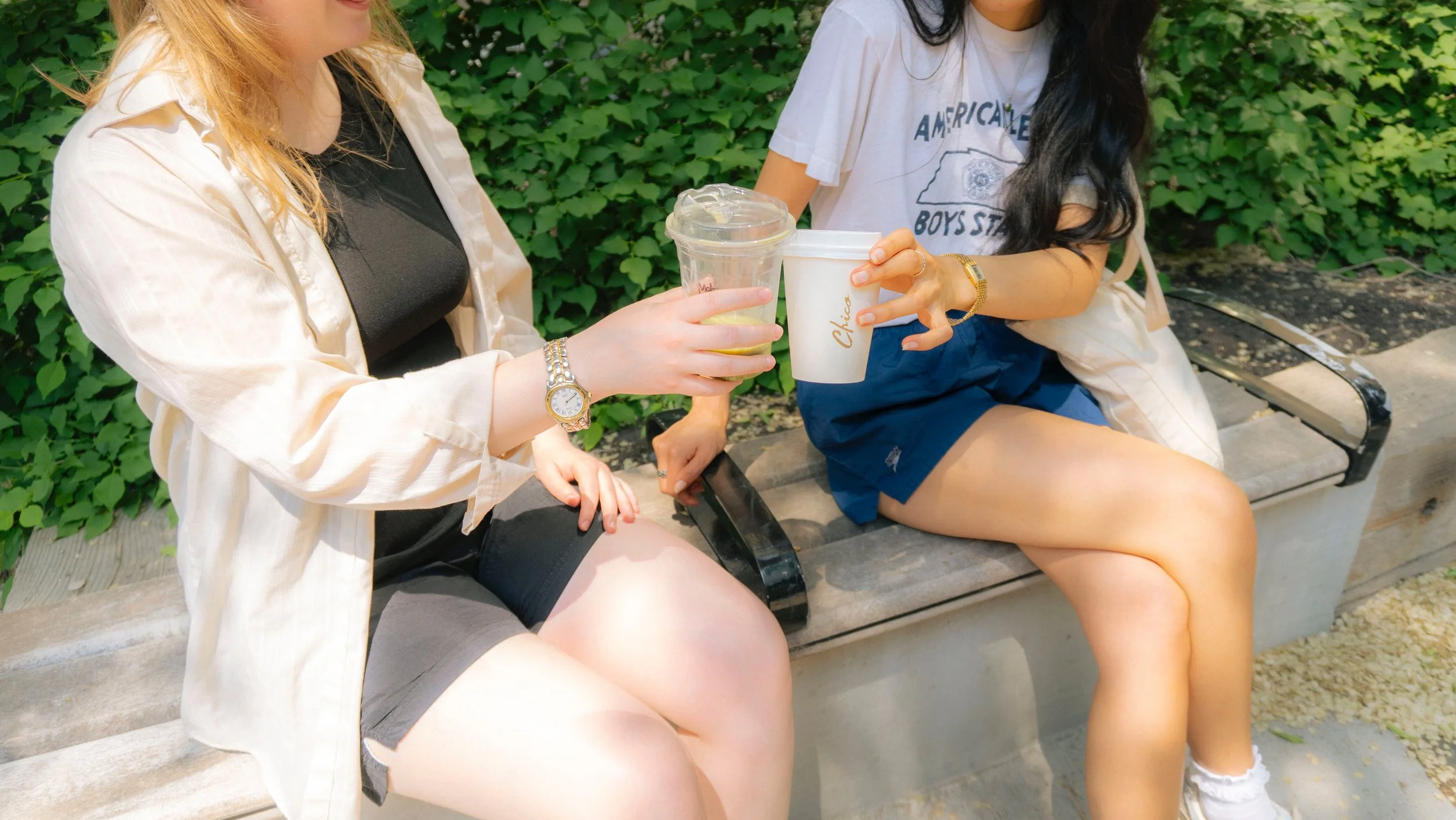 Two women sitting on a bench outdoors, holding drinks and engaging in conversation. One woman has blonde hair, wears a beige jacket over a black top, and a watch. The other woman has dark hair, wears a white T-shirt, blue shorts, and white sneakers.