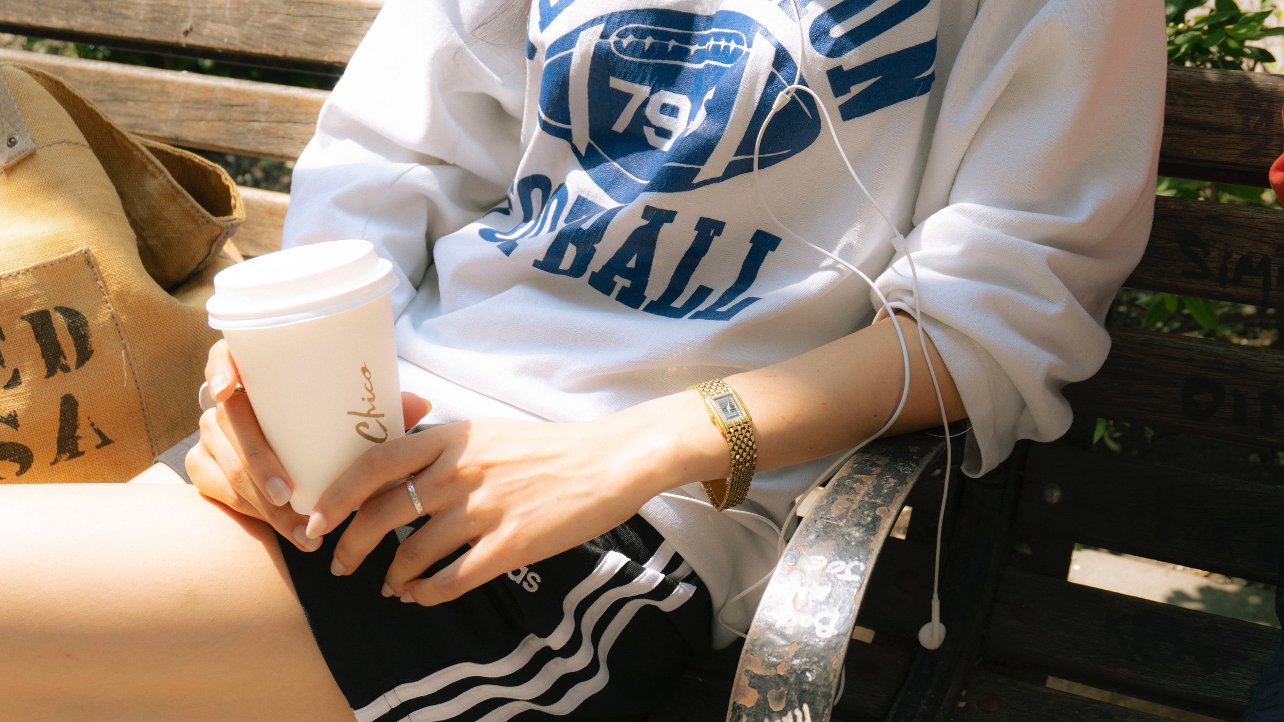 Person sitting on a wooden park bench, holding a disposable coffee cup, wearing a white sweatshirt with blue graphics, black shorts with white stripes, a gold watch, and a ring, with a tote bag beside them and earphones in.