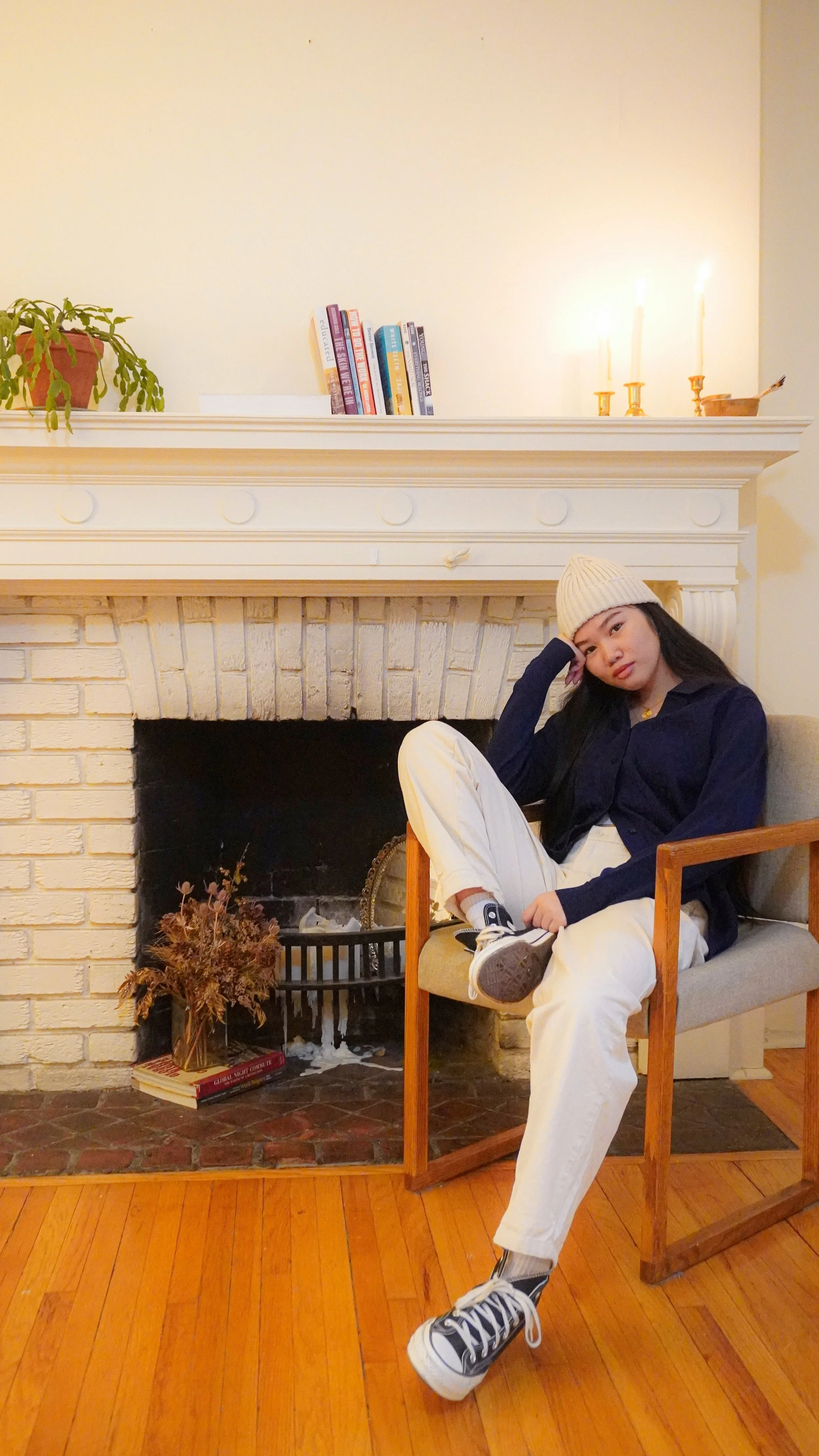 Young woman sitting in a beige armchair in front of a white brick fireplace, wearing a white knit hat, navy sweater, white pants, and black and white sneakers, with a relaxed pose and thoughtful expression.