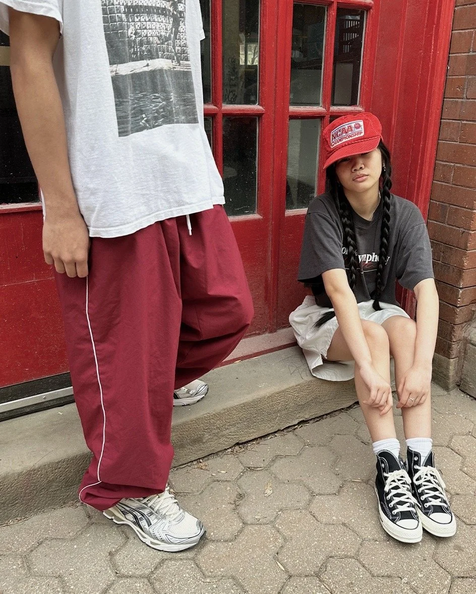 Two people in casual streetwear, one standing wearing a white graphic tee and red pants, the other sitting wearing a gray shirt, shorts, black high-top sneakers, and a red hat, in front of a red door.