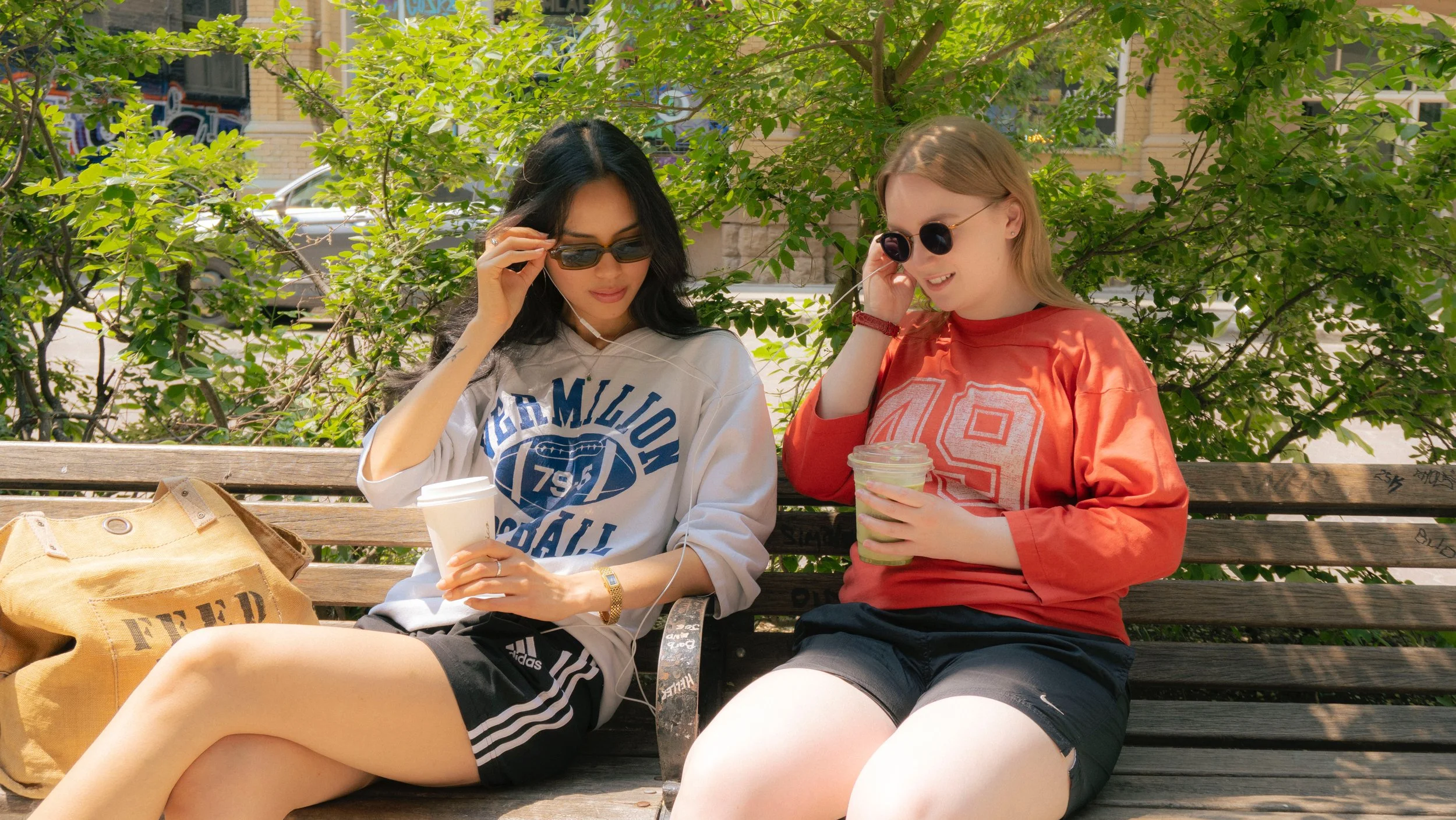 Two young women sitting on a wooden park bench, wearing casual athletic clothing and sunglasses, each holding a drink, with a greenery background and a brown bag labeled 'FUEL' beside them.