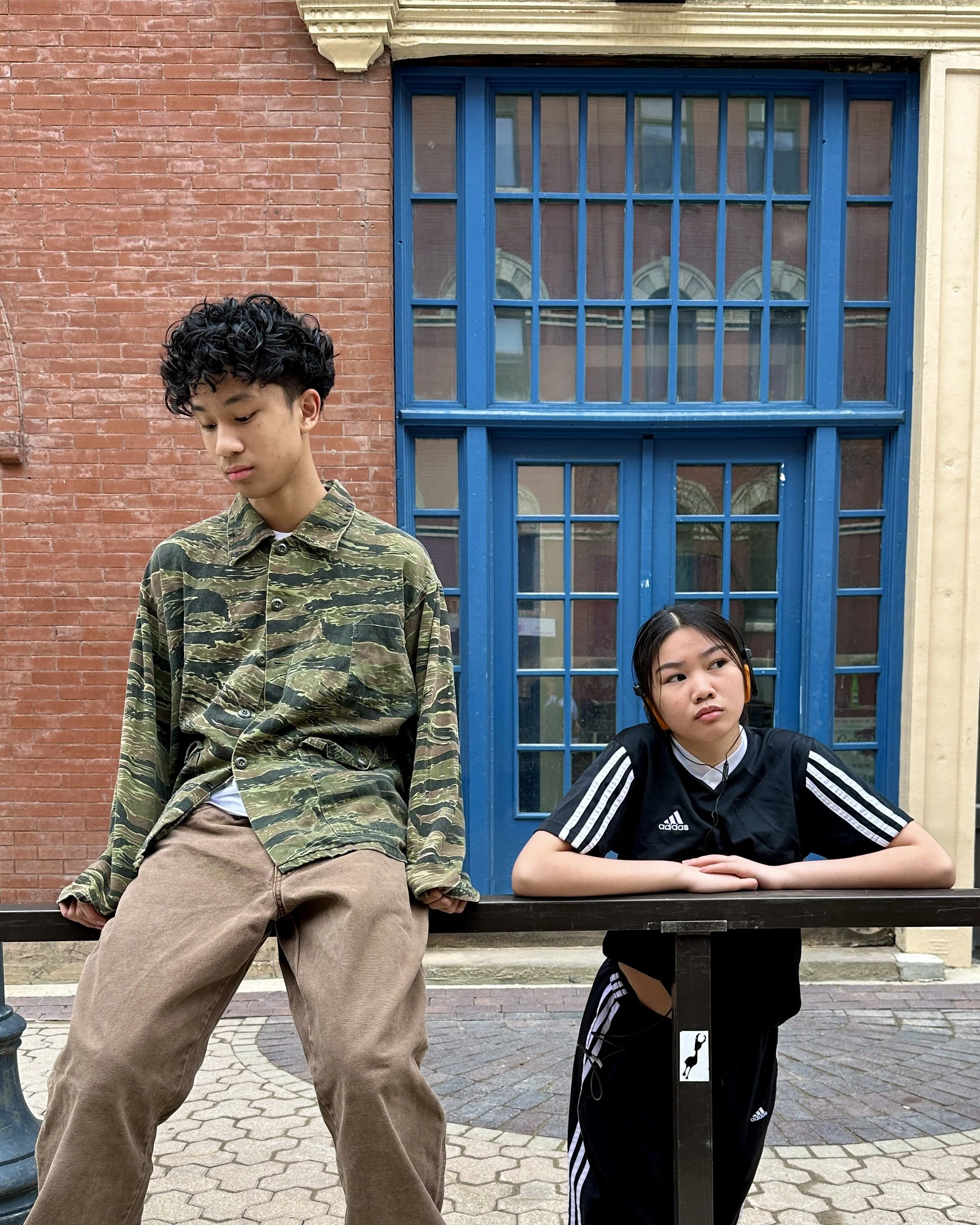 Two teenagers in front of a blue window, one wearing a camo shirt and the other in a black athletic outfit with white stripes.