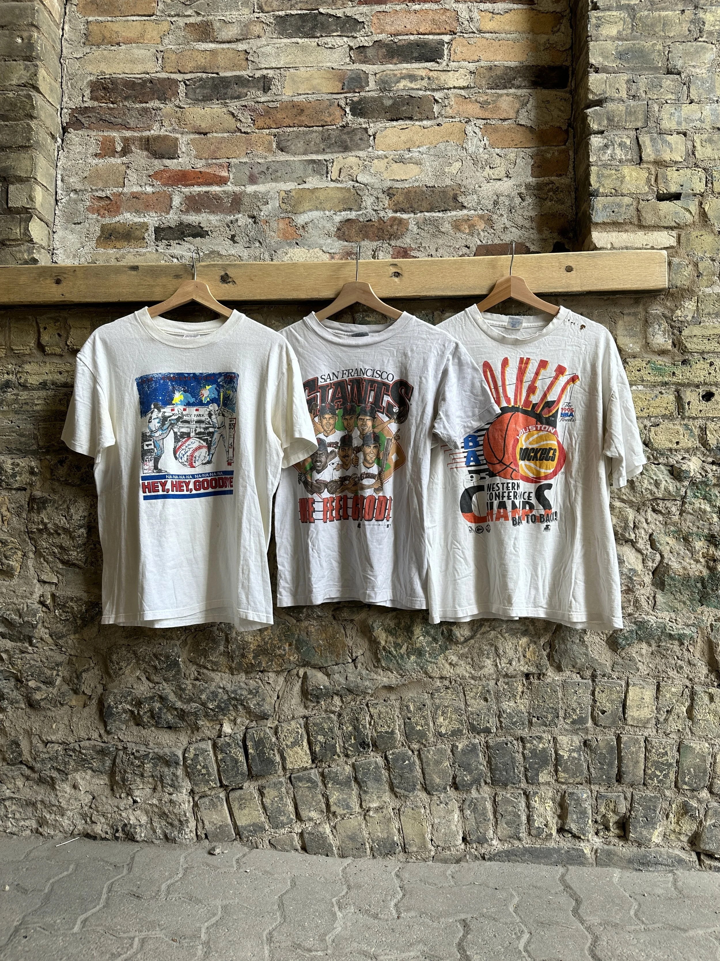 Three vintage graphic T-shirts on wooden hangers against a brick wall.