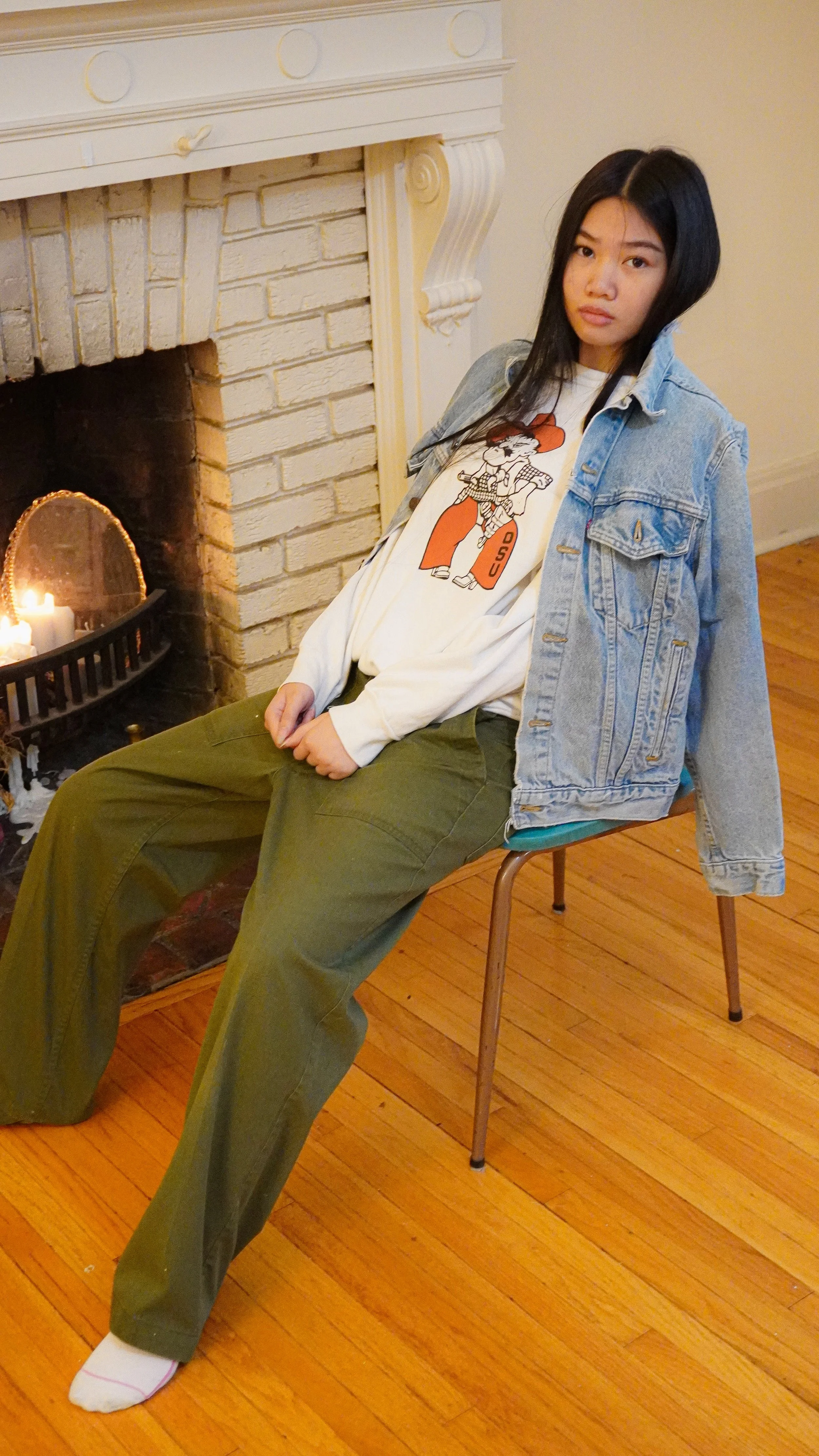 Girl sitting on a chair near a fireplace, wearing a denim jacket, white graphic t-shirt, olive green pants, and white sock.