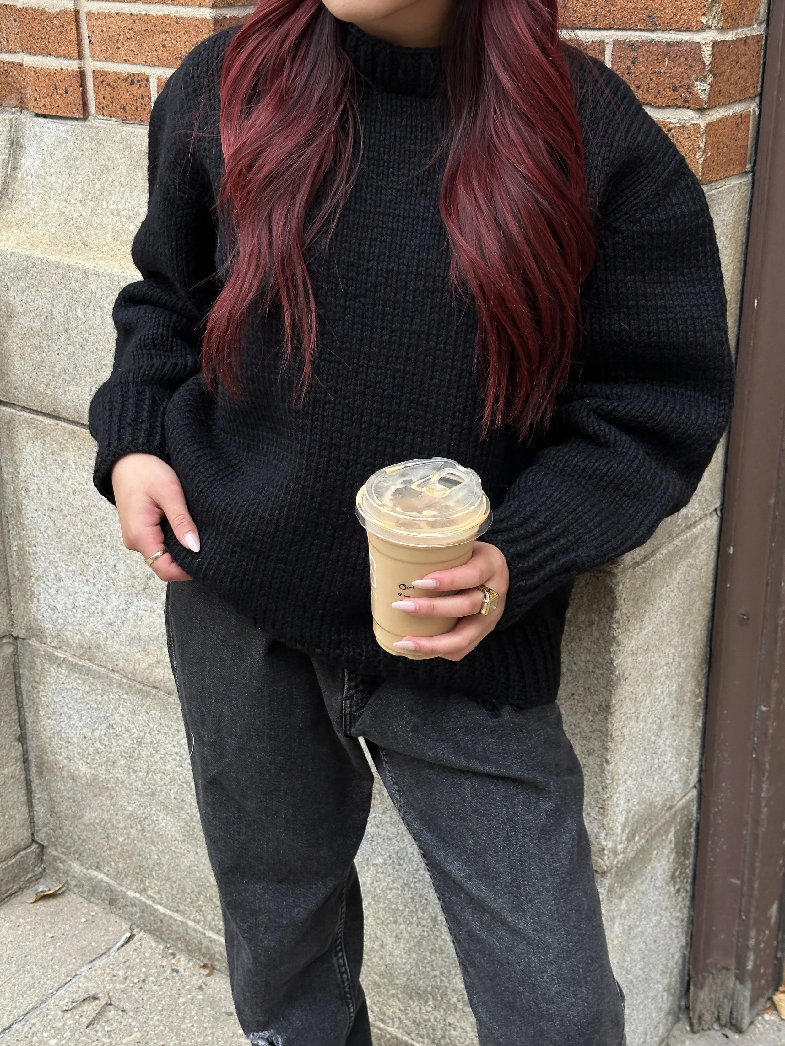 Person with long red hair wearing a black sweater and jeans holding an iced coffee.