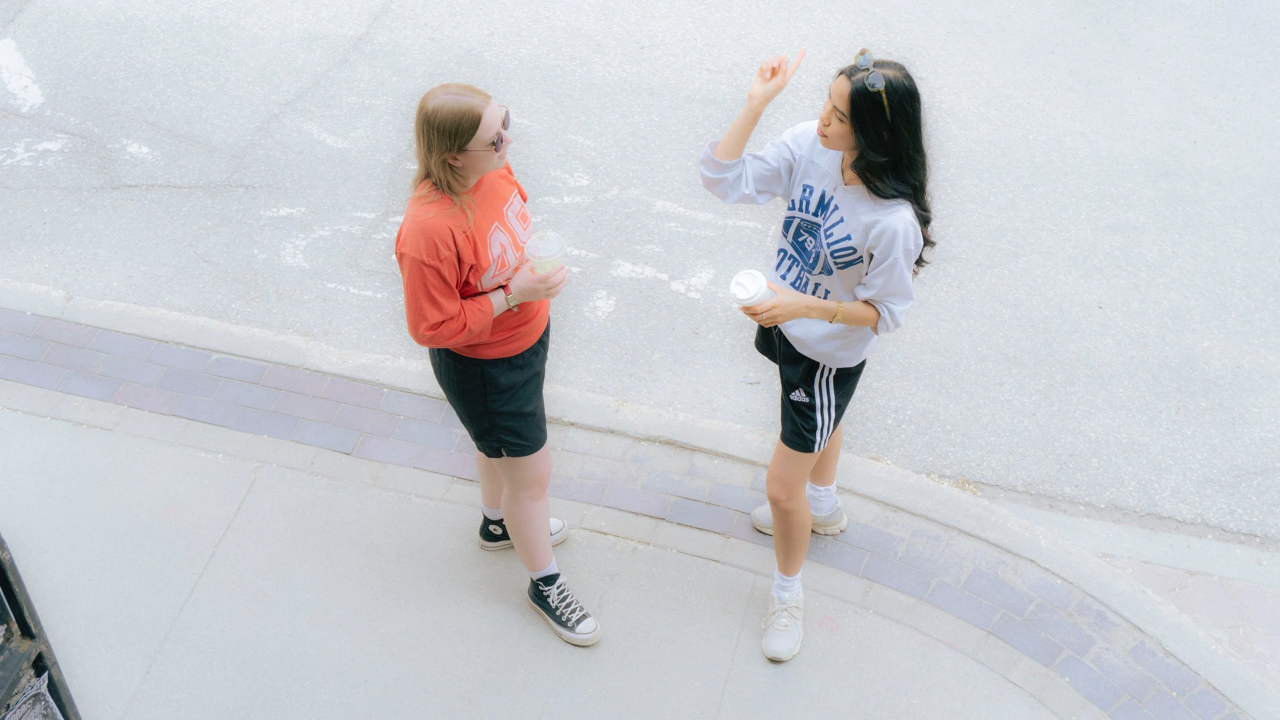 Two young women standing on a sidewalk talking and holding drinks, viewed from above.