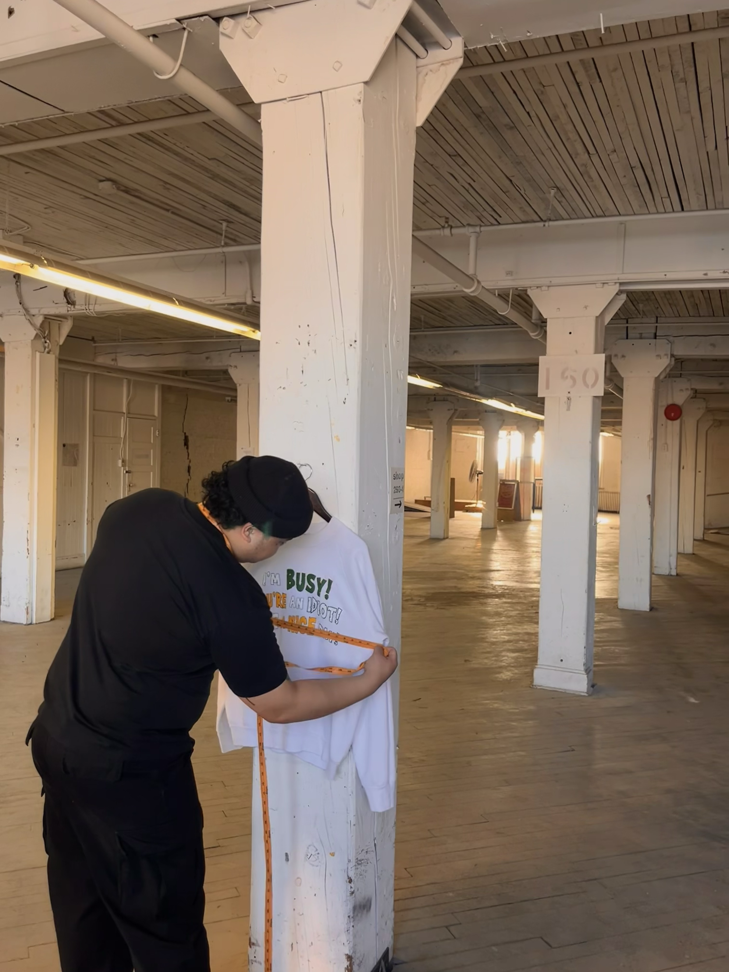 Person measuring a white sweatshirt with text in a large, industrial-style room with columns and wooden floors.