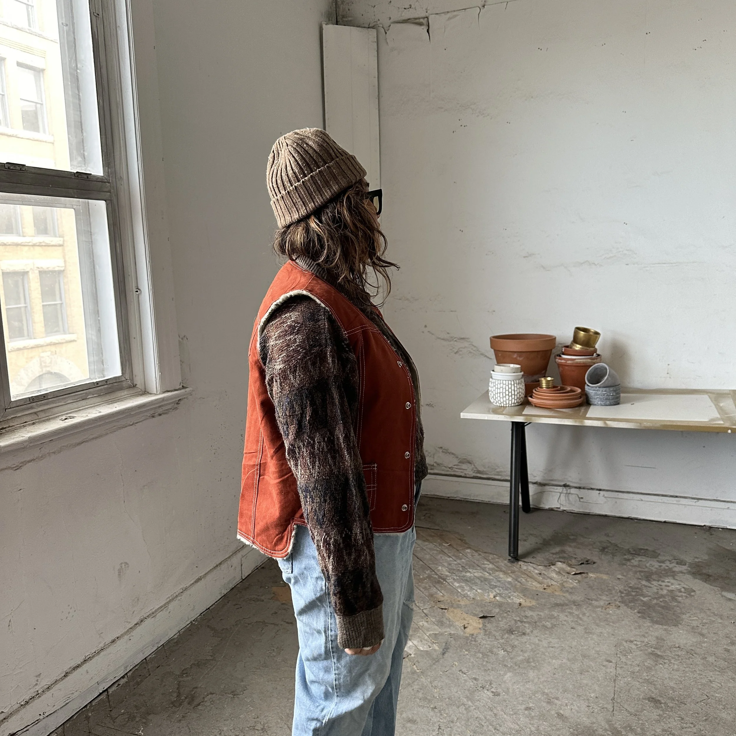 Person wearing a beanie, brown vest, and patterned sweater stands in a rustic room with a table holding various pottery items.