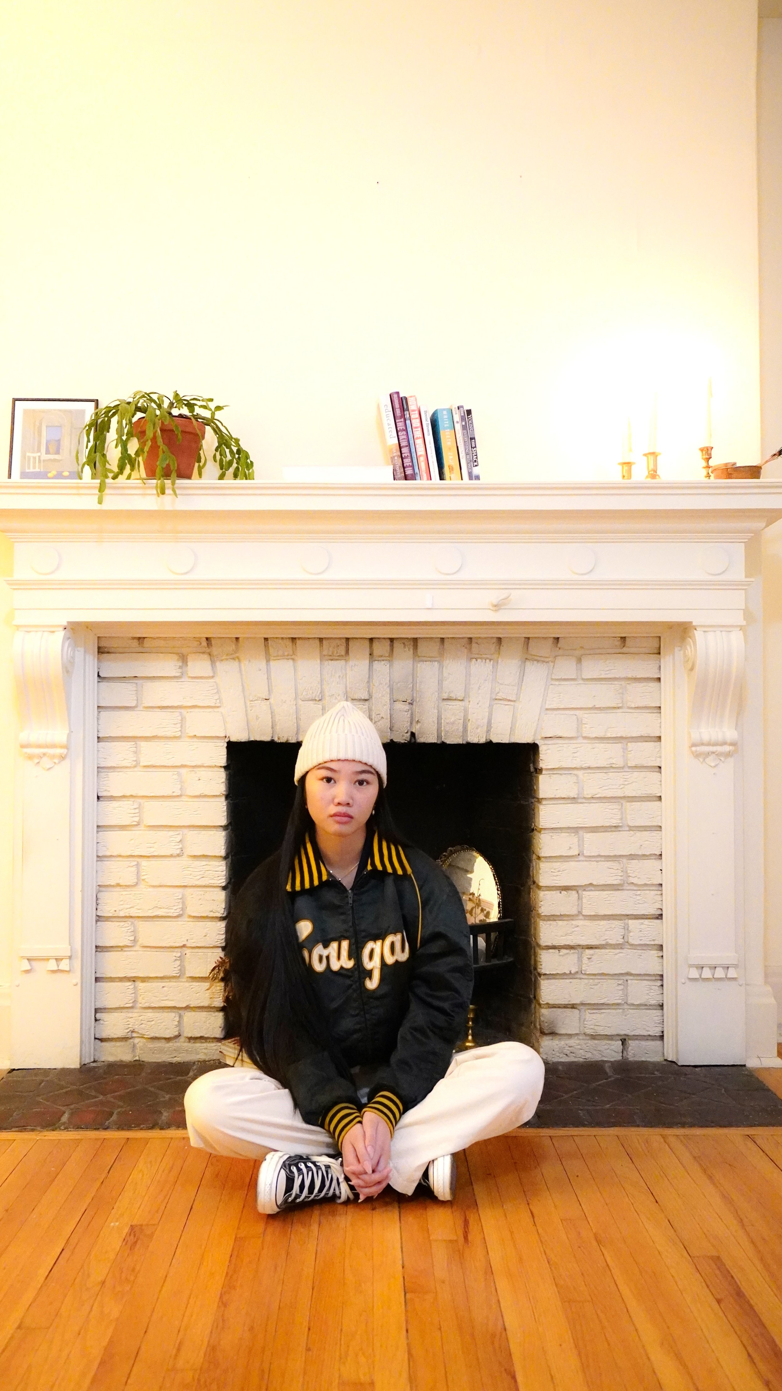 Young woman with long black hair, wearing a white beanie, black and yellow jacket, white pants, and black sneakers, sitting cross-legged on a wooden floor in front of a white brick fireplace in a living room.