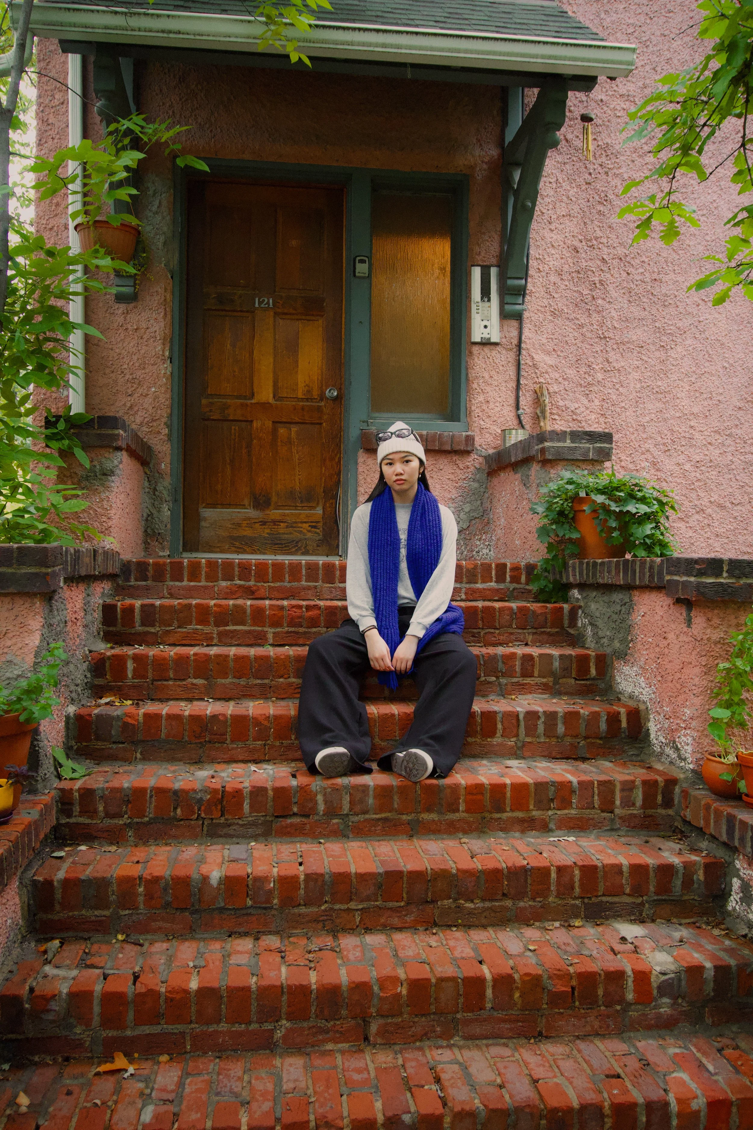 A young woman sits on brick steps outside a pink stucco house with a wooden door, potted plants, and foliage around the entrance.