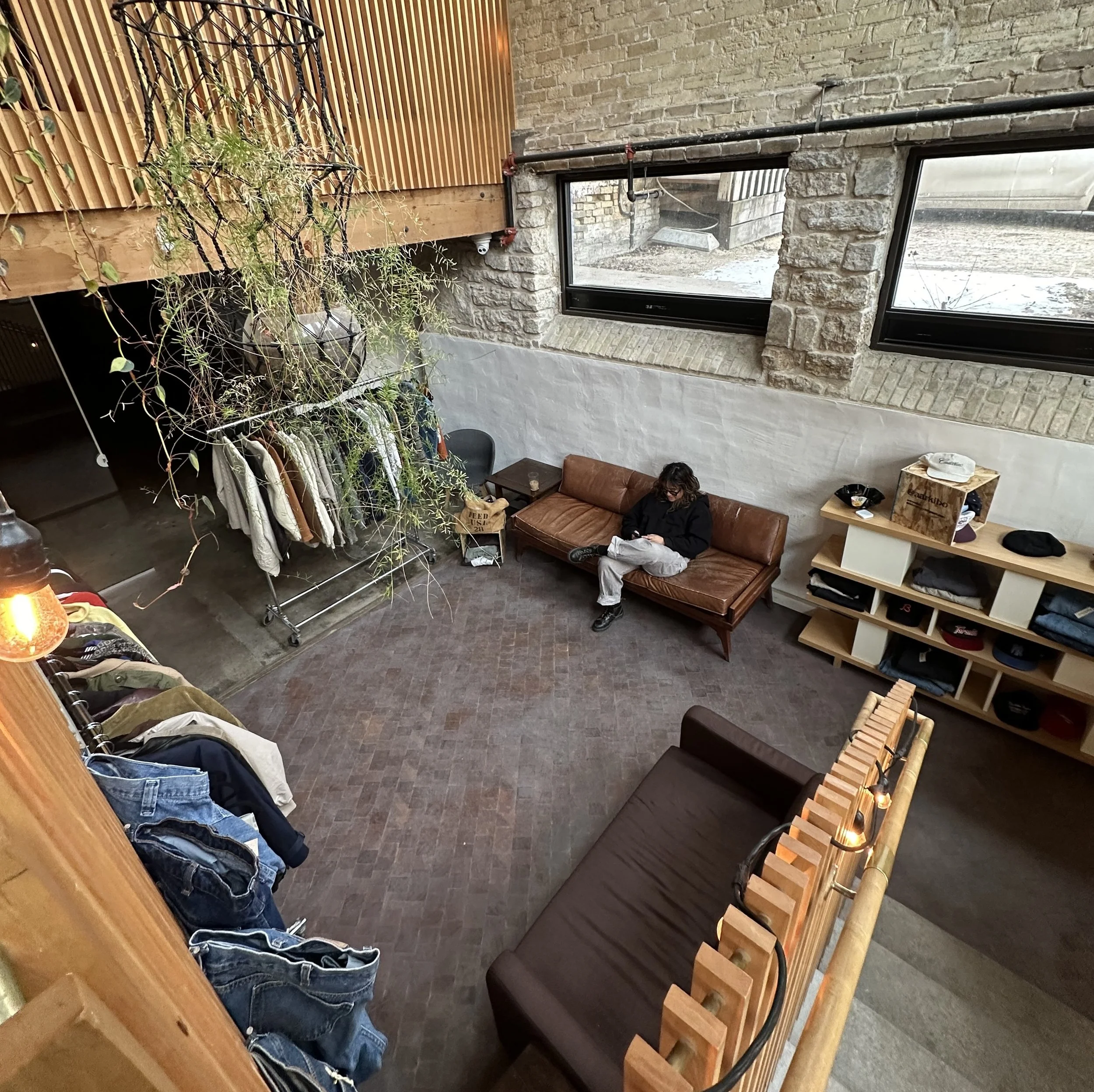 Industrial-style living space with brick walls, leather sofa, and clothing rack. Person sitting on sofa, and plants hanging from ceiling. Natural light from large windows.