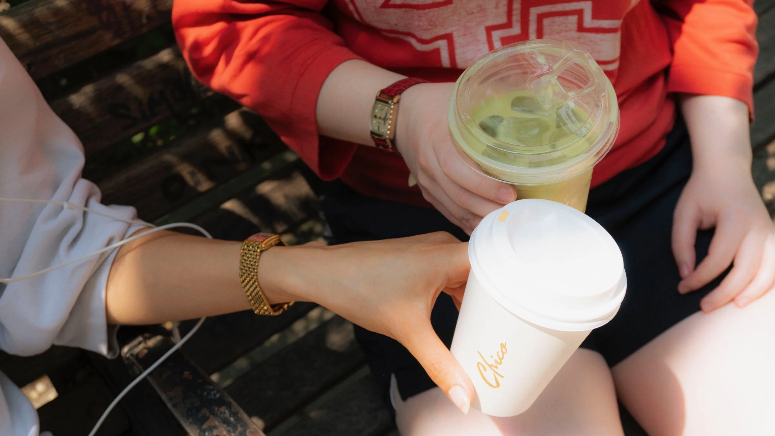 Two people sitting outdoors sharing drinks, one holding a white to-go coffee cup and the other holding a large clear cup with a yellow beverage and ice, on a wooden bench.