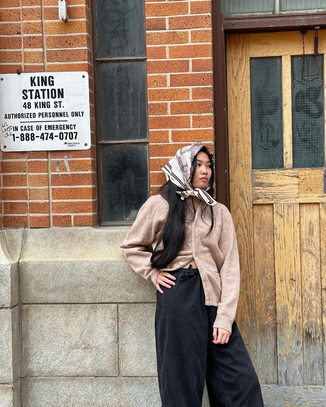 Person leaning against a brick building near a "King Station" sign, wearing a headscarf, beige cardigan, and black pants.