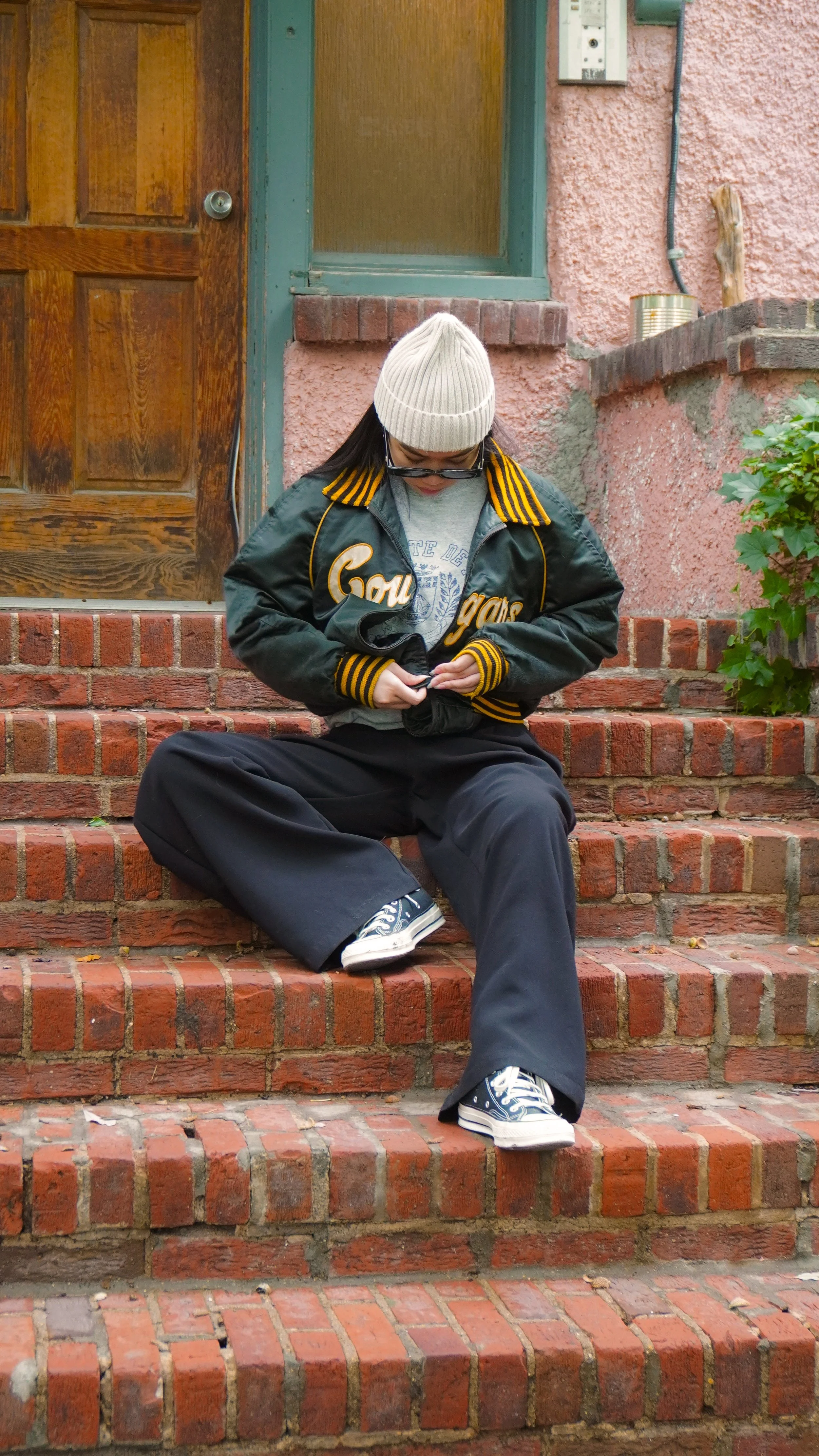 A person dressed in a black and yellow varsity jacket, wide pants, sneakers, a gray t-shirt, a beige knit hat, and glasses sitting on brick steps outside a house, looking down and adjusting their clothing.
