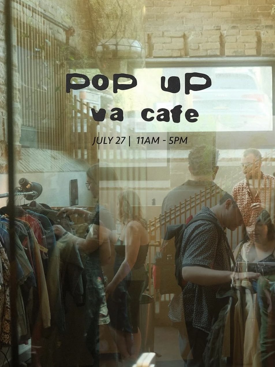 View through a window showing people browsing clothes at a pop-up event with text on the glass reading "pop up ya cafe, July 27, 11AM - 5PM."