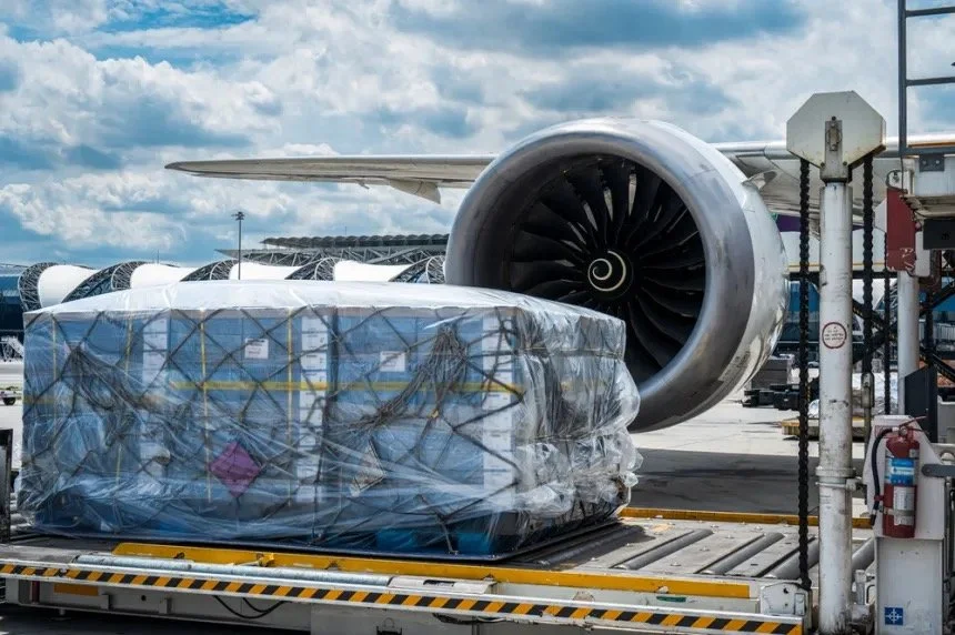 Cargo wrapped in plastic on a loading platform near an airplane engine at an airport.