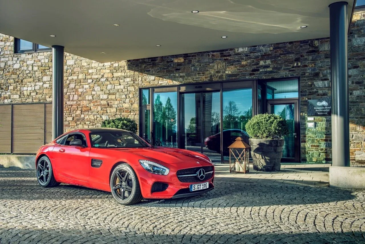 Red Mercedes-Benz sports car parked in front of a building with stone facade and large glass doors, under a covered area.