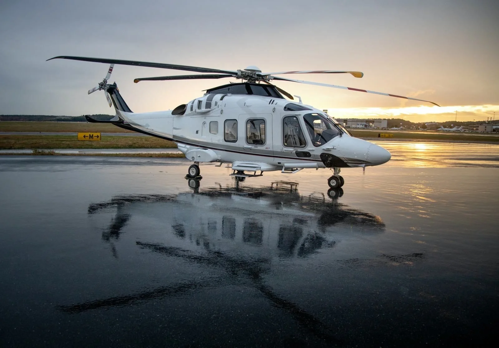 A white helicopter parked on a wet tarmac during sunset, with reflections visible on the water surface.