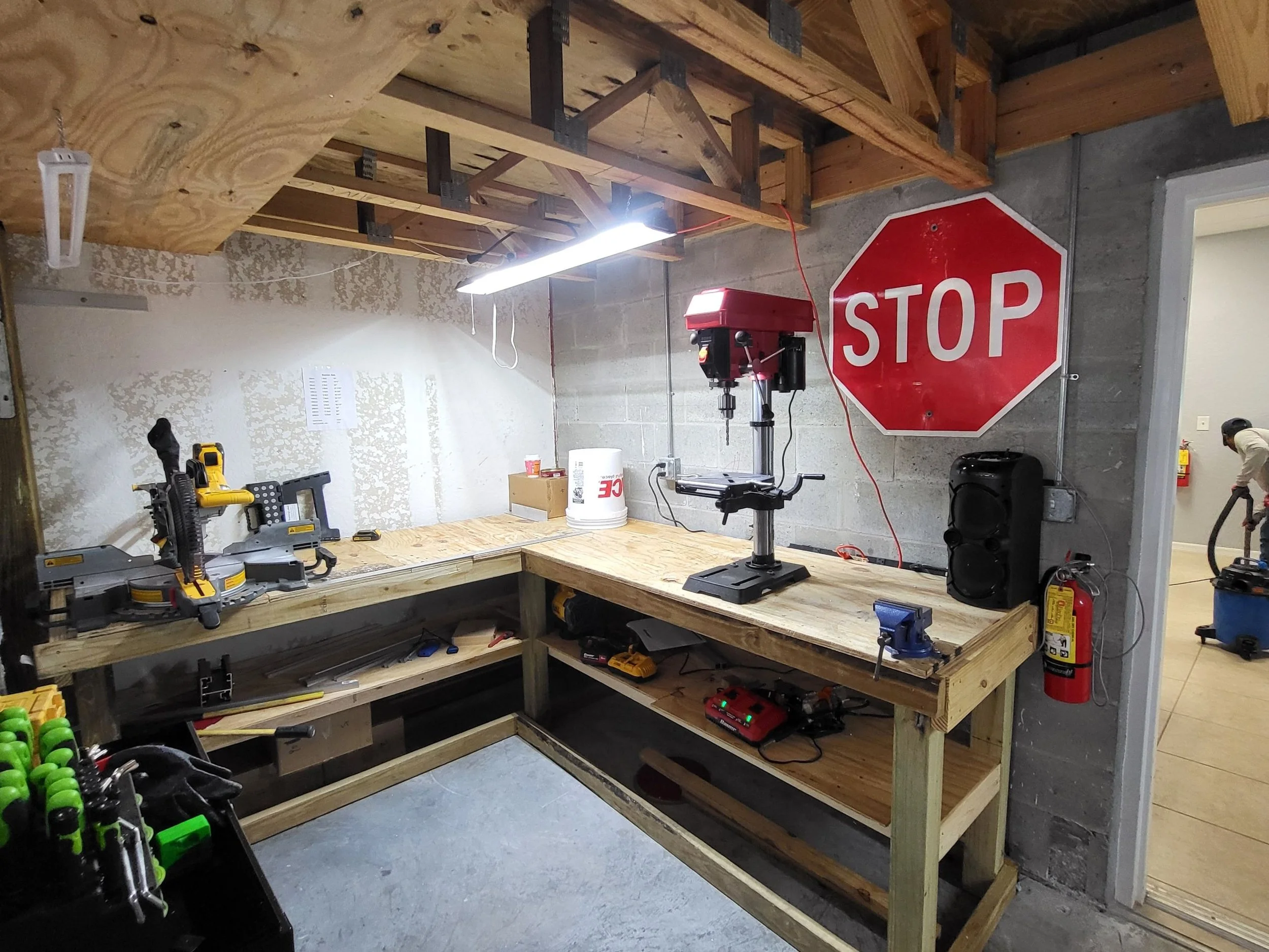 A workshop with a large workbench made of unfinished wood, various power tools, a red stop sign, and construction materials, with a cinderblock wall and a person working in the background.
