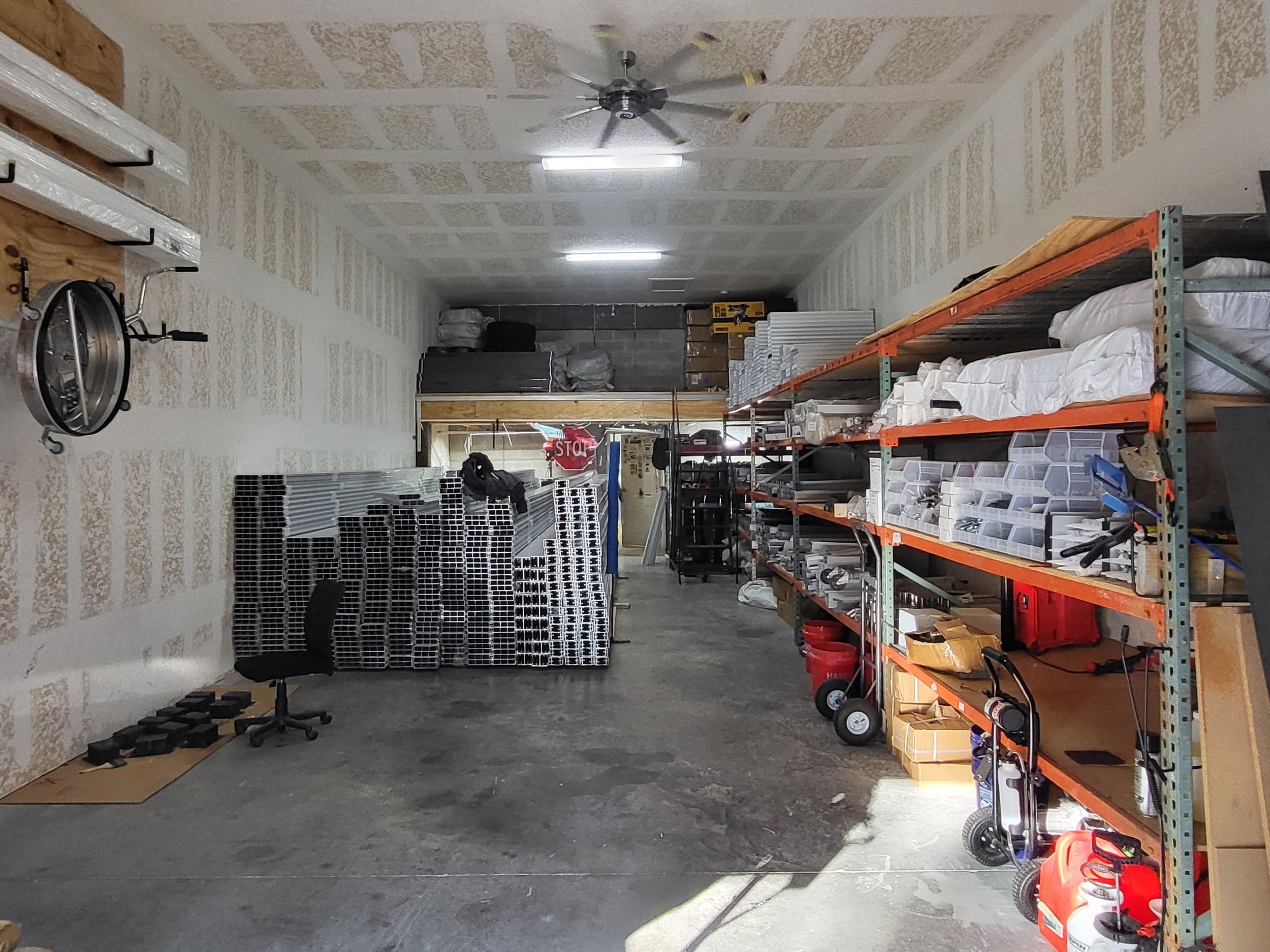 A storage room with shelves holding various tools and supplies, a ceiling fan, fluorescent lights, and a concrete floor.