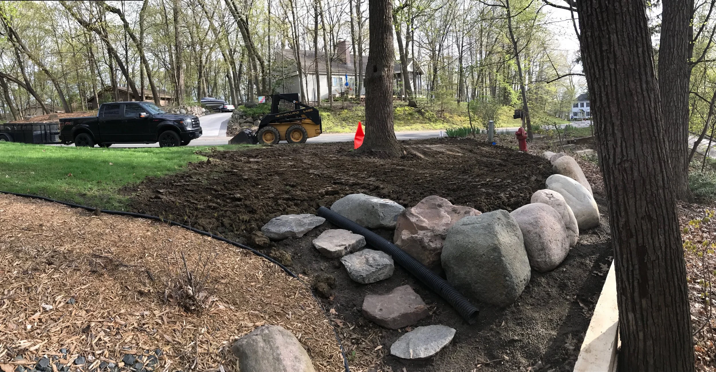 A yard under landscaping or construction, with soil prepared for planting or other work, bordered by large rocks and trees, with construction equipment, a vehicle, and a house visible in the background.