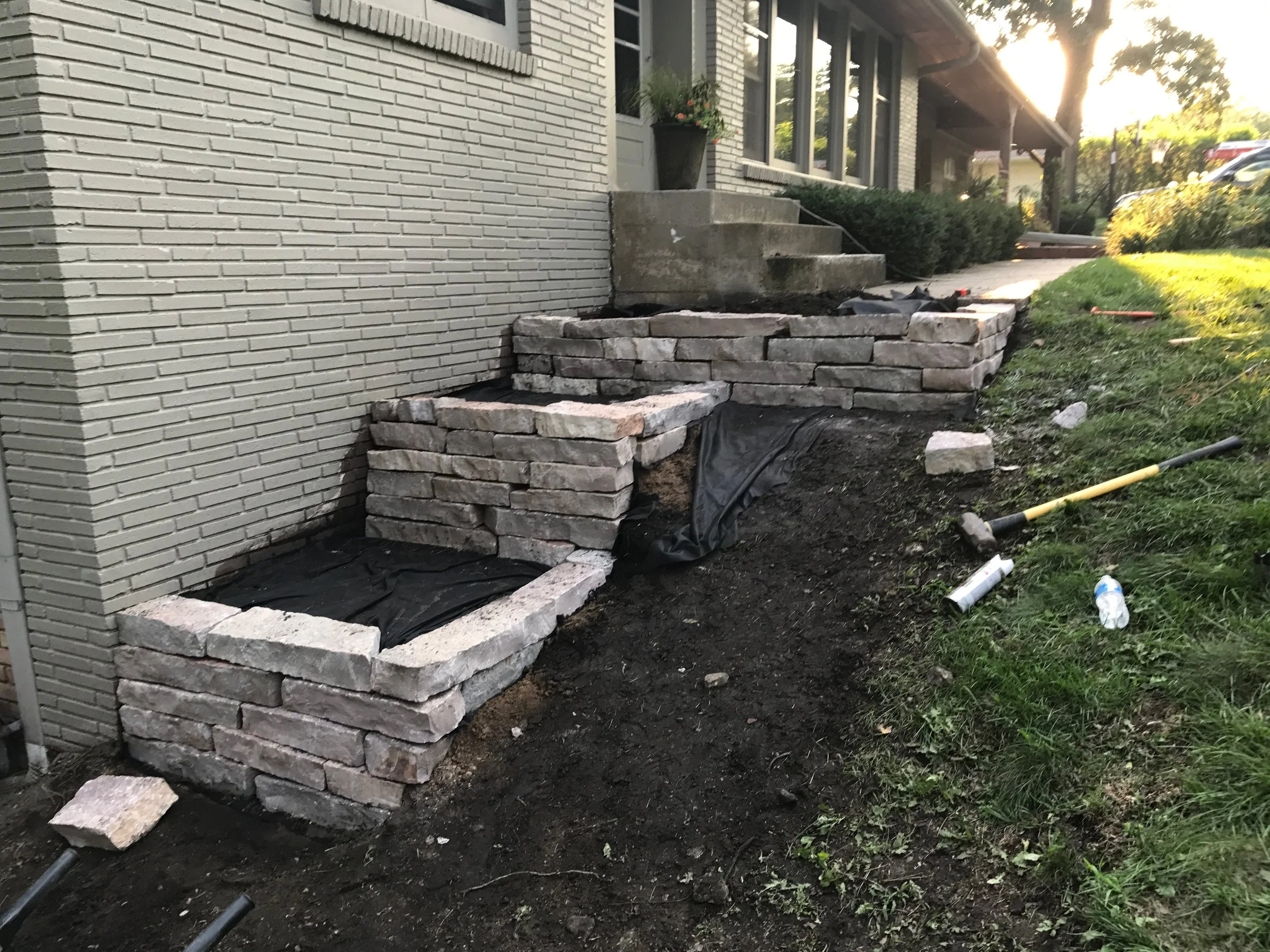 Building exterior with a brick wall, stone steps, and a garden area under construction with landscaping materials nearby.