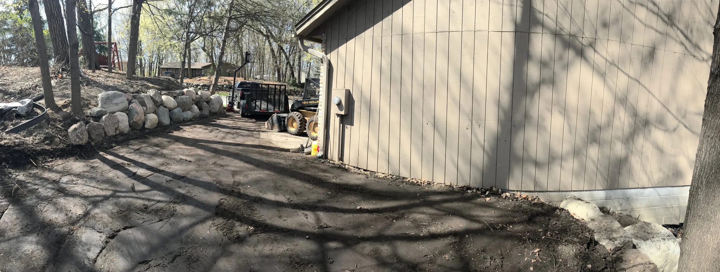 A backyard with a dirt pathway, rocks lining one side, and construction equipment including a small loader and trailer parked near a beige wooden building.