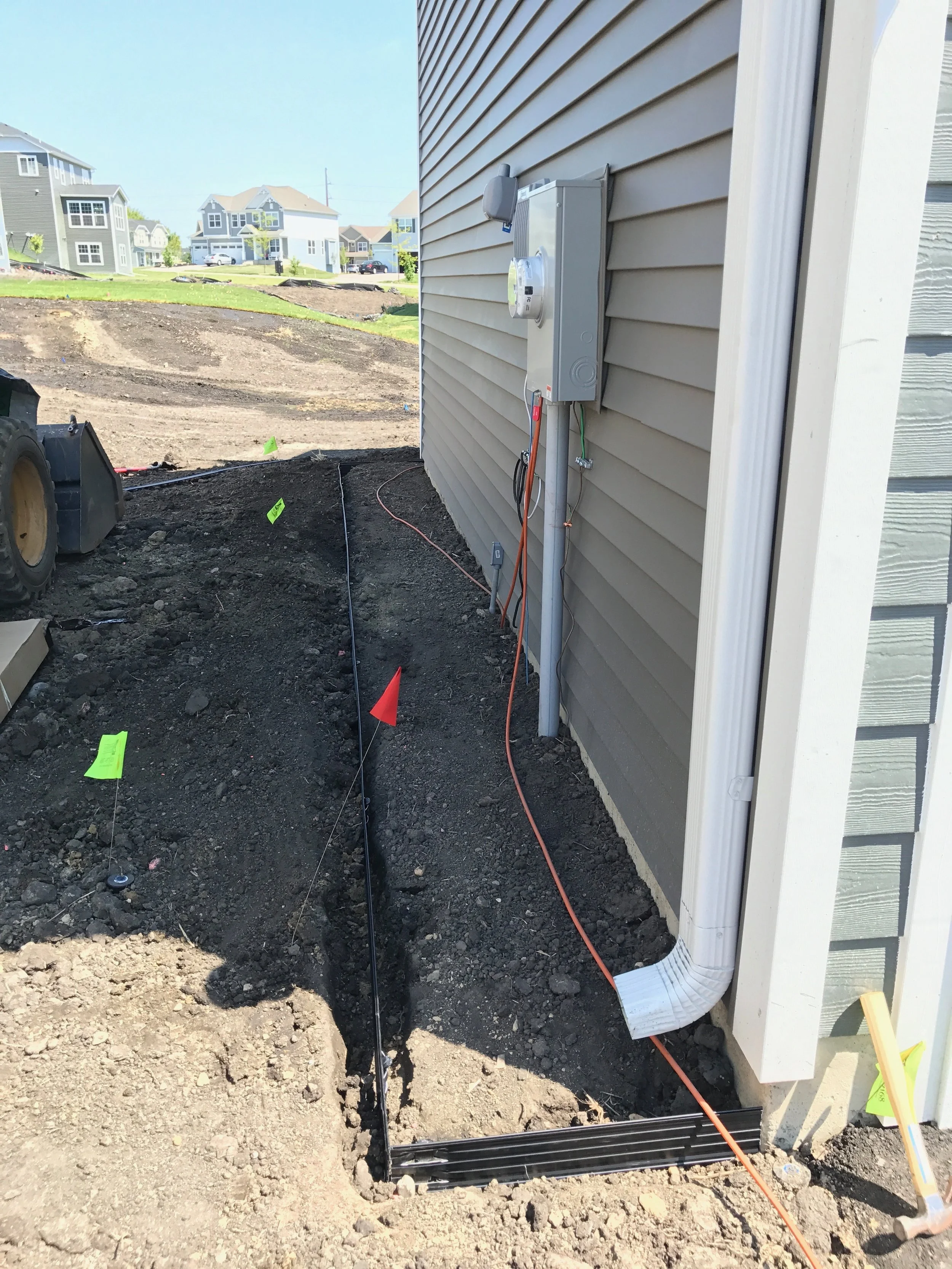 Construction site with electrical box, exposed wiring, and dirt trench along the side of a house with beige vinyl siding.
