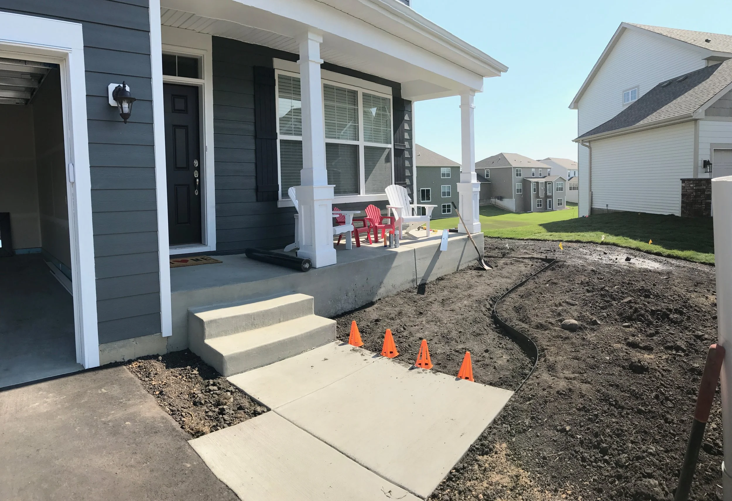 A house with a dark gray exterior and white trim, front porch with three chairs, some construction cones, and ongoing landscaping work on the yard.