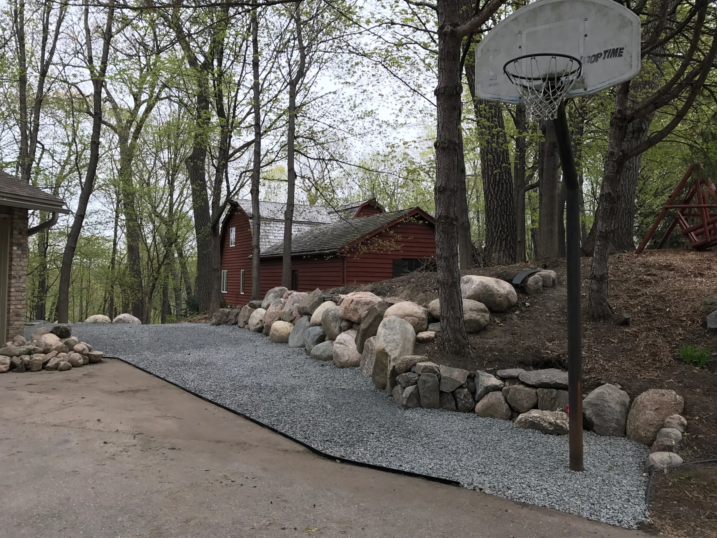 A gravel pathway next to a house, surrounded by large rocks and trees, with a basketball hoop in the yard.