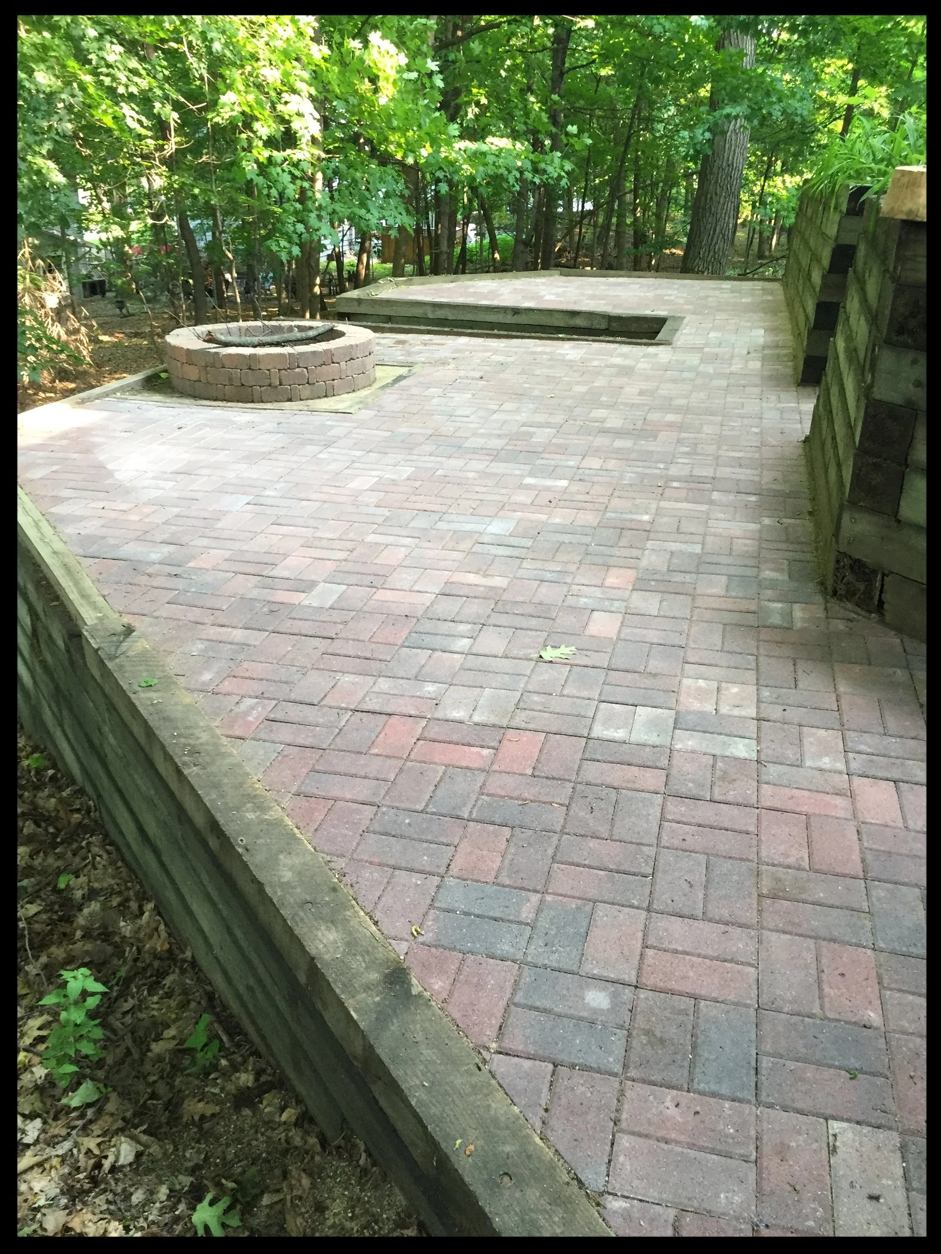 A paved outdoor patio with brick pavers, featuring a round fire pit made of bricks and a rectangular sunken area, surrounded by green trees and a wooden retaining wall.