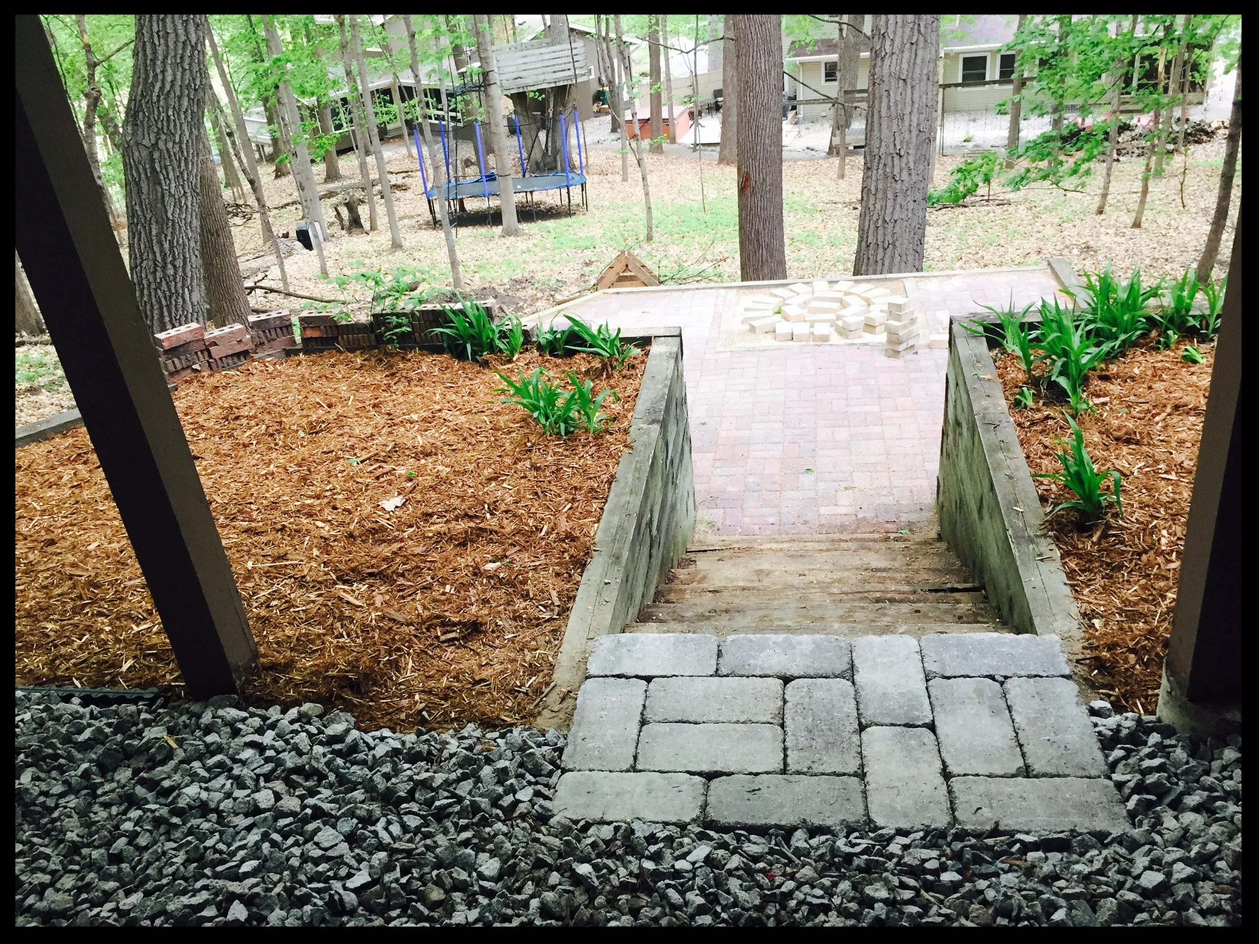 View of a backyard with a small set of stairs leading from a porch to a backyard paved with bricks, with a garden bed on each side filled with soil and some green plants, trees in the background, and a trampoline and outdoor playhouse in the backyard