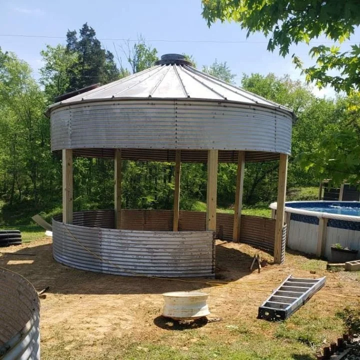 A partially constructed round metal gazebo in a backyard with trees, an above-ground pool, and outdoor equipment.