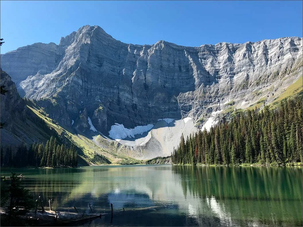 photo of a mountain surrounded by pine trees and a lake in the summer. There is snow on the mountains.