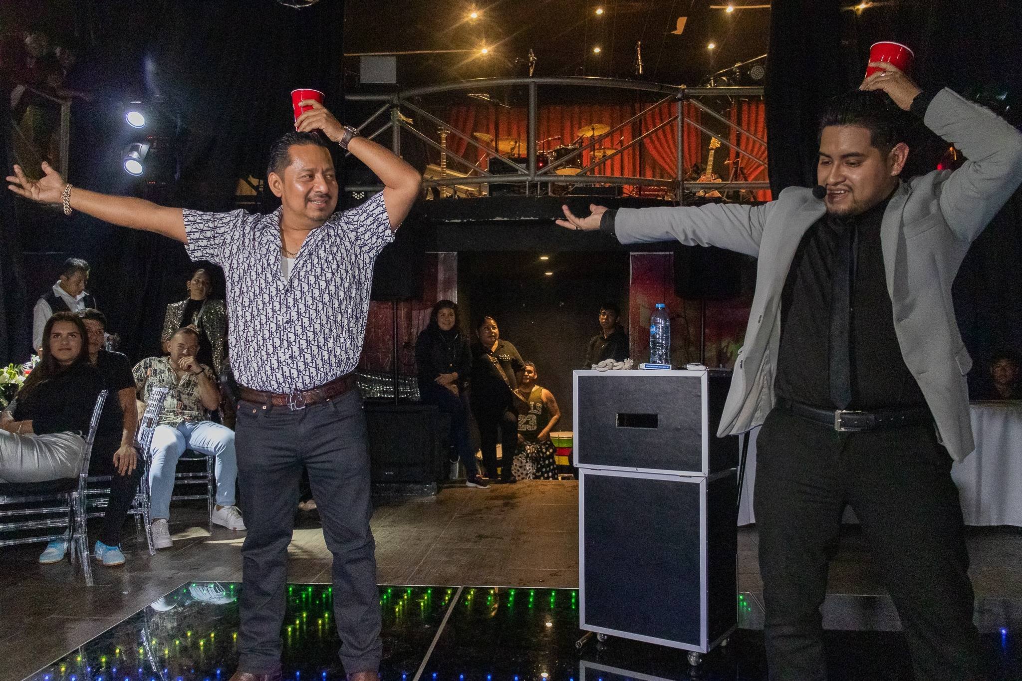 Dos hombres en un karaoke con vasos rojos en la cabeza y el público observando.