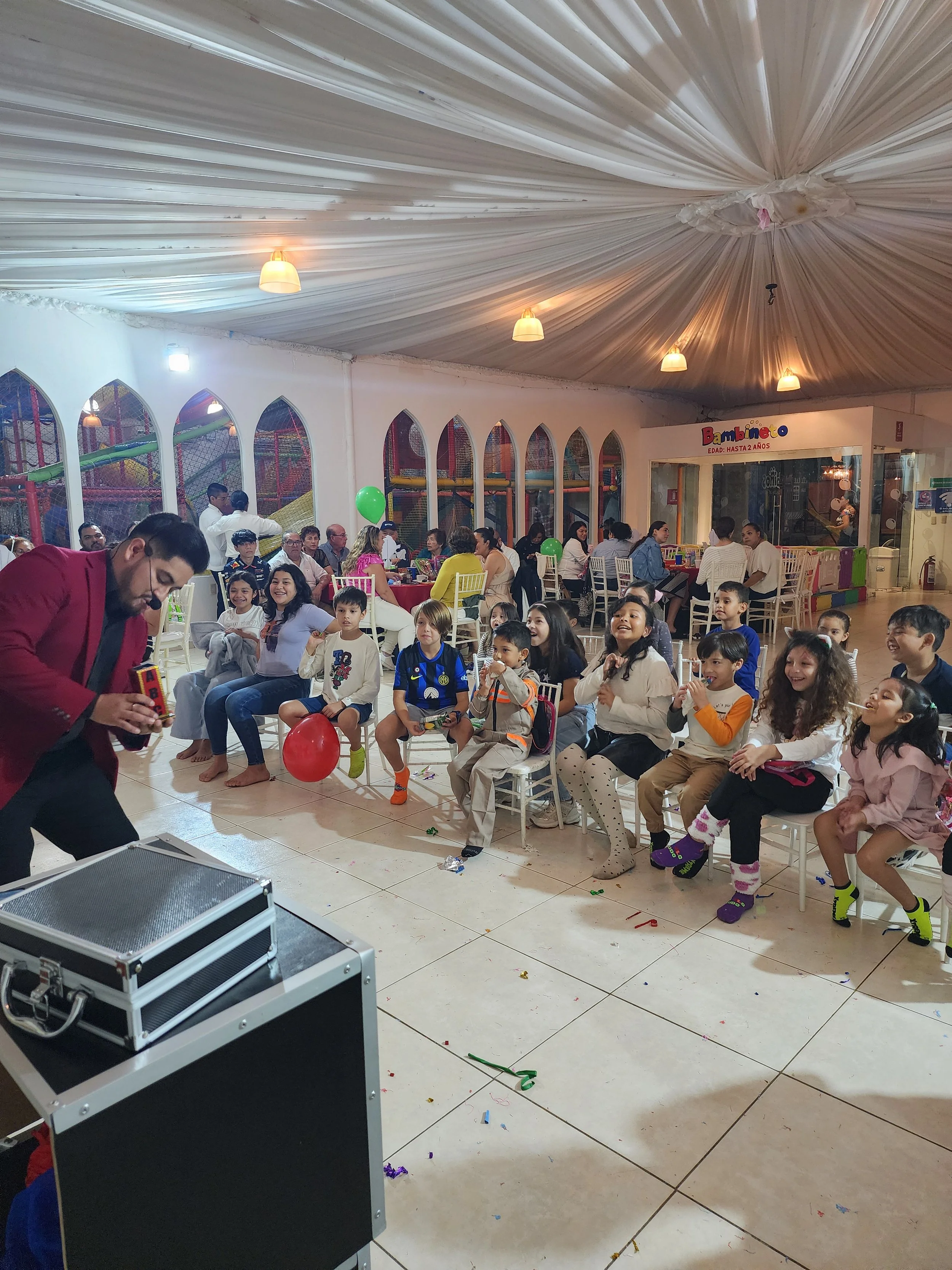 Celebración de fiesta infantil con un mago actuando frente a un grupo de niños y adultos, rodeados de globos y decoraciones festivas en un salón.