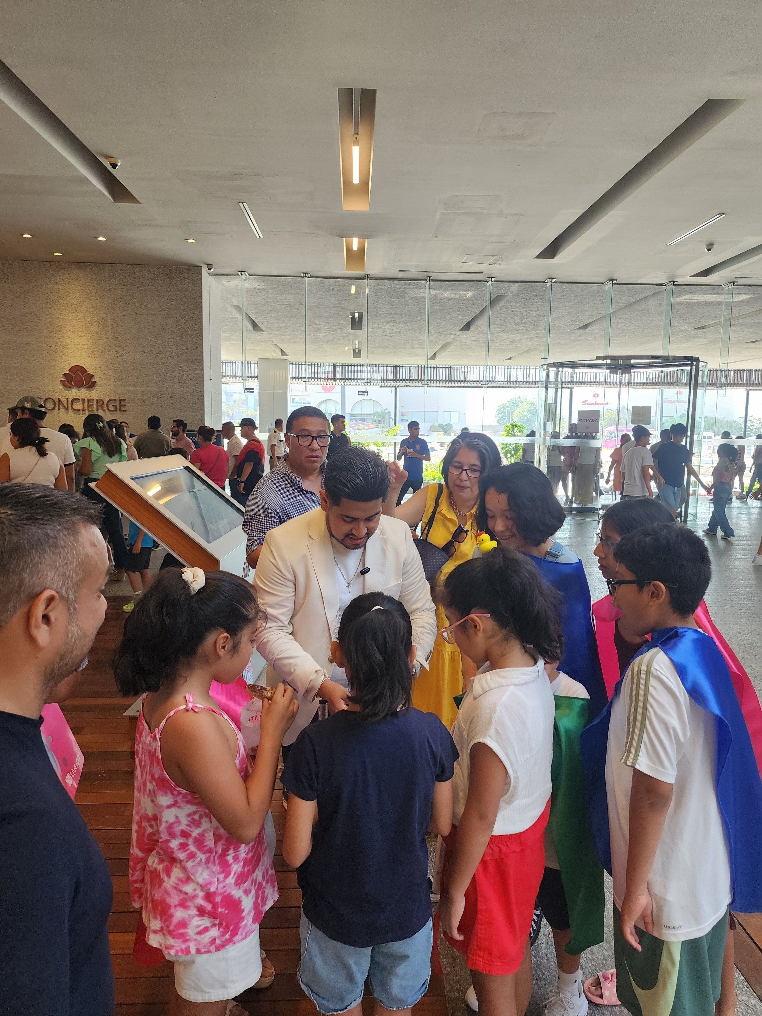 Grupo de personas, principalmente niños, rodeando a un hombre vestido de blanco en un vestíbulo concurrido.