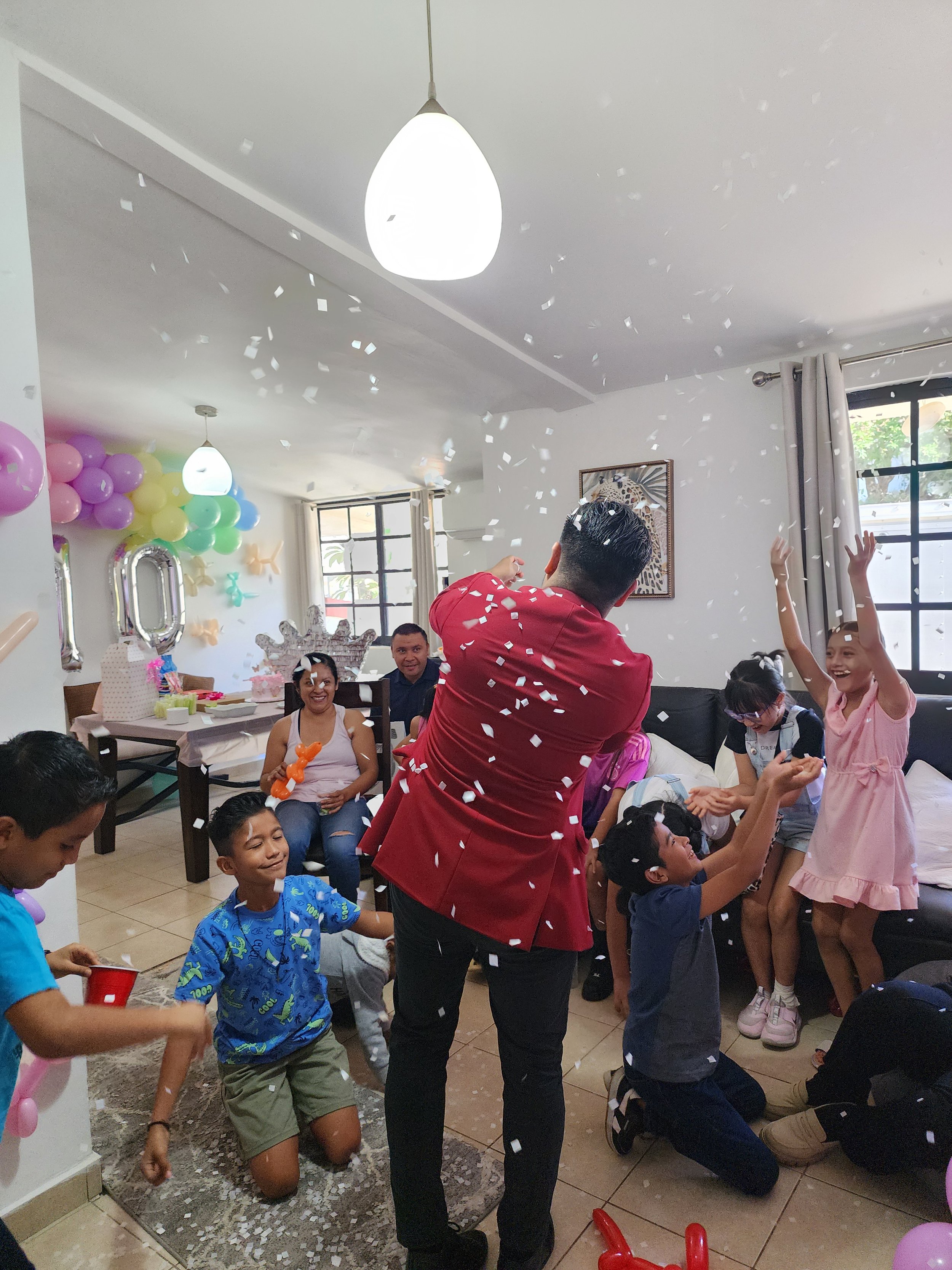 Celebración de cumpleaños infantil con niños y un animador lanzando confeti en una sala decorada con globos y números de cumpleaños.