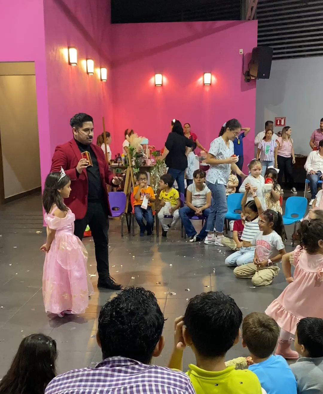 Fiesta infantil en un salón con paredes rosadas, donde hay un animador realizando un espectáculo para varios niños sentados. Una niña vestida de princesa está de pie al frente. Varias personas están en el fondo, charlando y sirviendo comida.