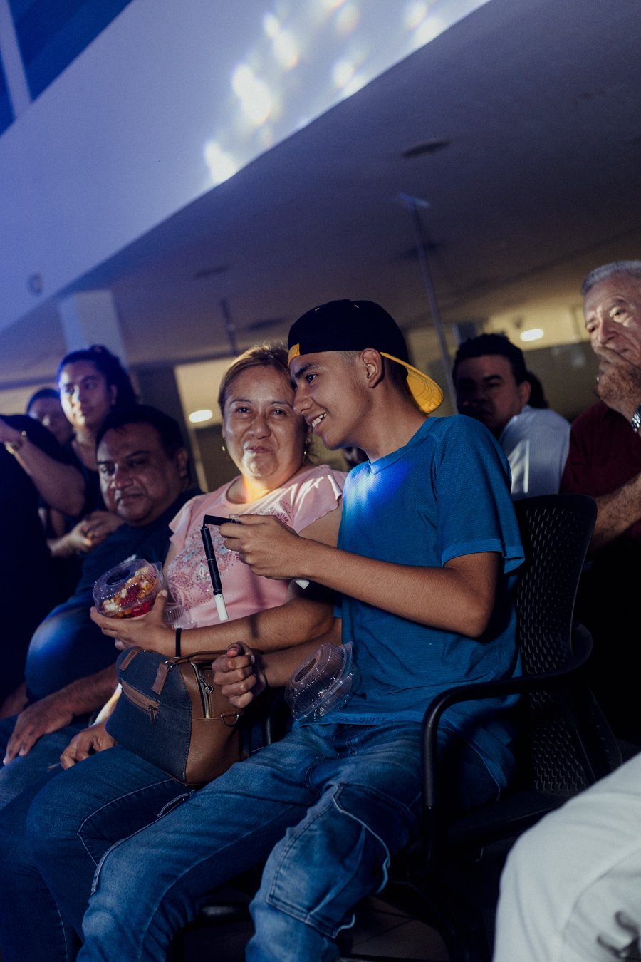 Grupo de personas sentadas, algunas sonriendo, en un ambiente iluminado. Un joven lleva gorra y camiseta azul, sosteniendo un dispositivo. Persona a su lado sostiene un recipiente con comida.