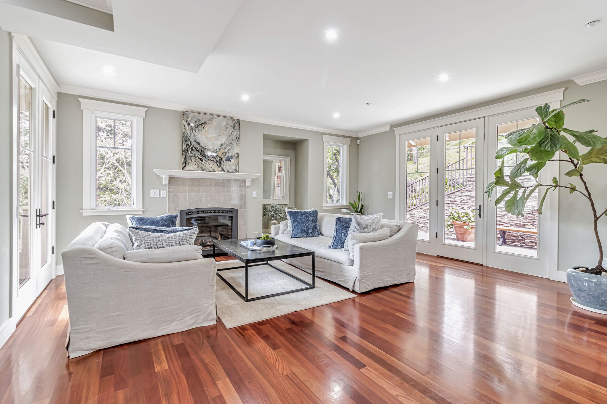 Bright living room with hardwood floor, white sofas, a glass coffee table, a fireplace with artwork above, potted plant, and French doors leading outside.