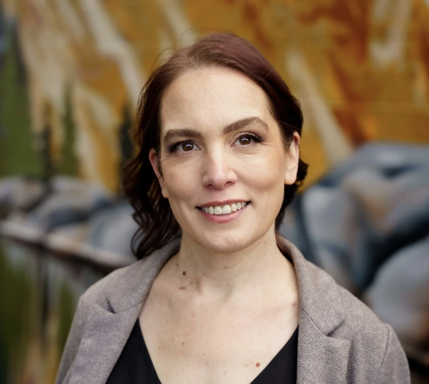 Smiling woman with wavy hair wearing a lavender cardigan, sitting in front of colorful abstract paintings.