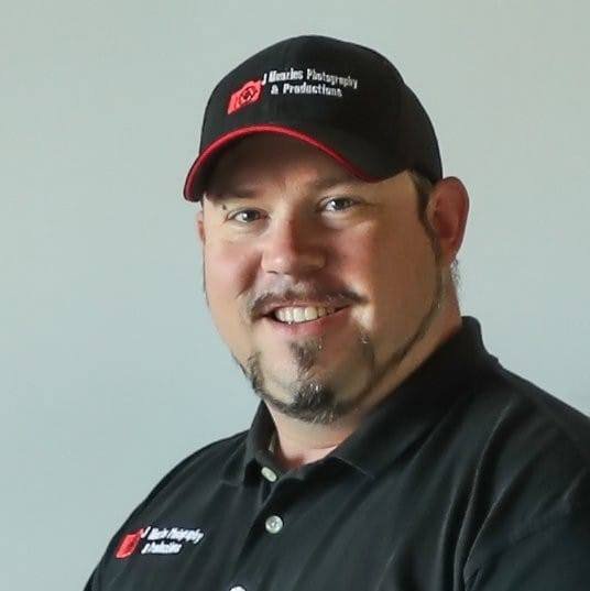 A man wearing a black baseball cap with red trim and logo, smiling against a plain background.