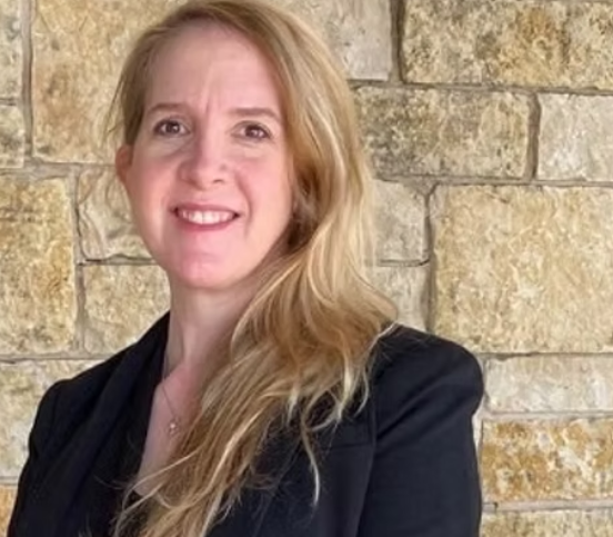 A woman with long blonde hair, smiling, wearing a black blazer, standing in front of a beige brick wall.