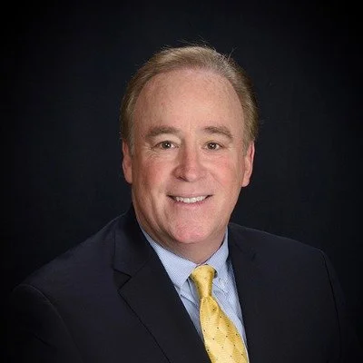 Professional headshot of a smiling man in a dark suit, light blue shirt, and yellow tie against a dark background.