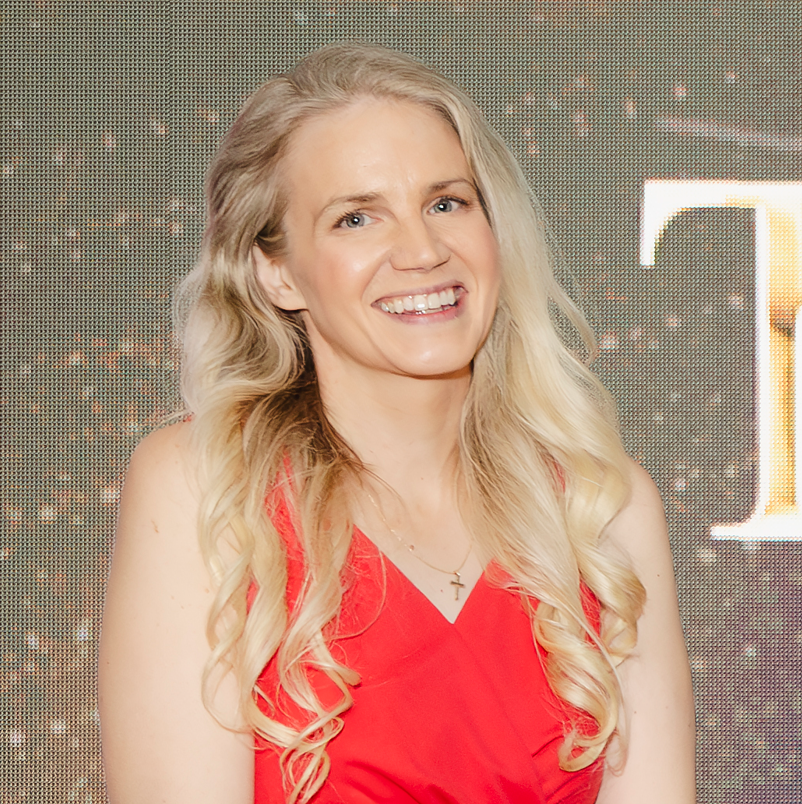 A smiling woman with long blonde hair standing outdoors during sunset with dark clouds in the background.