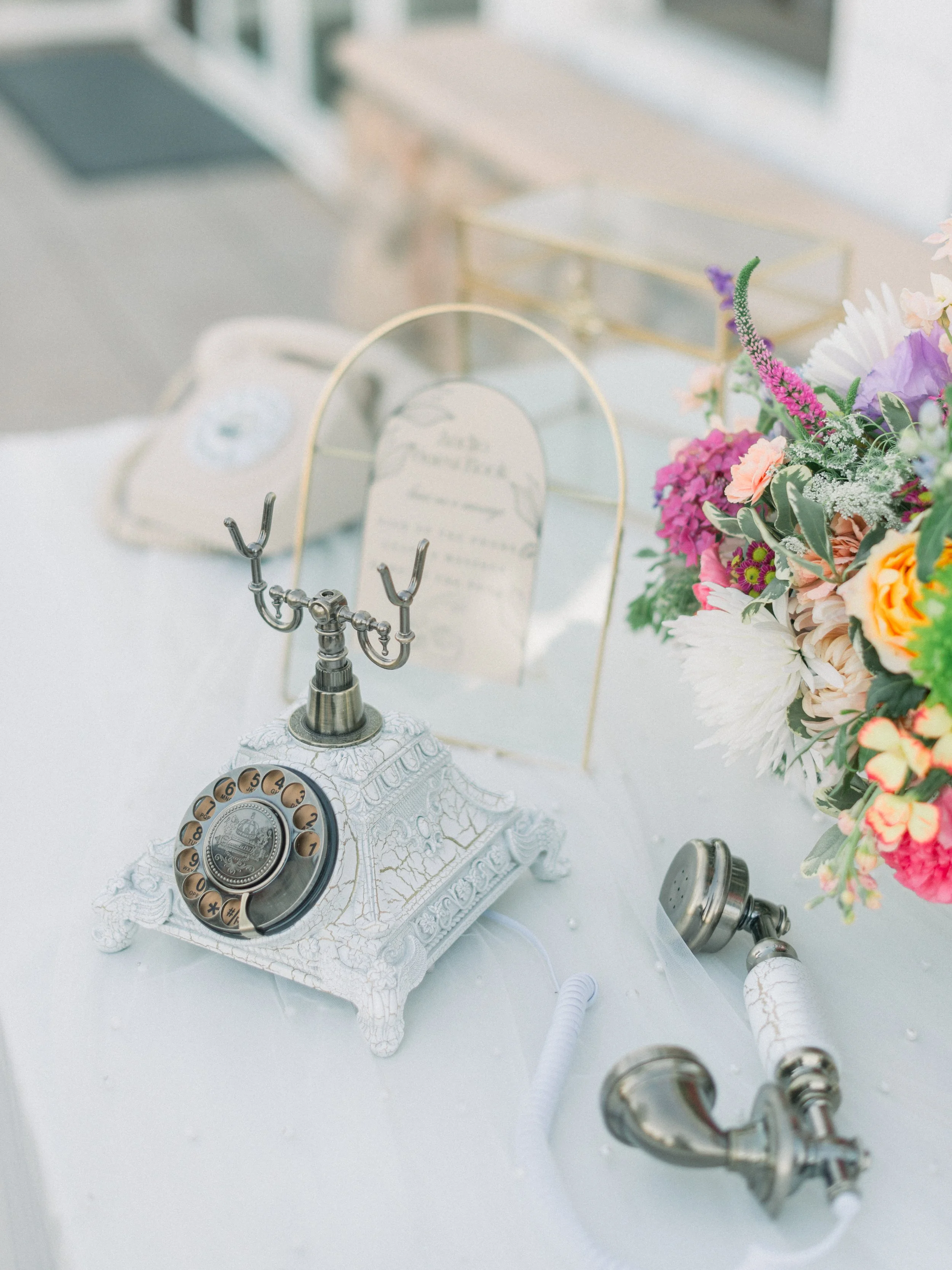 Vintage rotary dial telephone with ornate base on a table, alongside a colorful bouquet of flowers and a decorative card stand.
