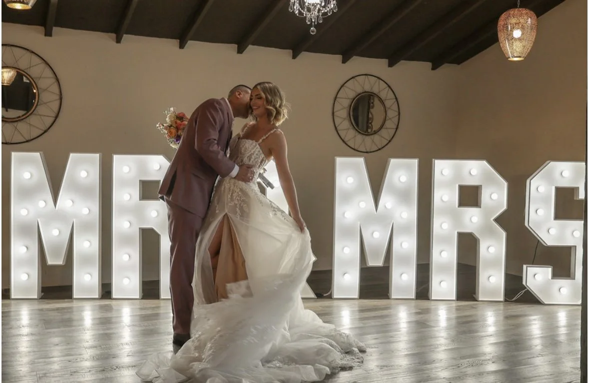 A bride and groom pose in front of illuminated 'MR & MRS' letters, with the bride holding a bouquet and lifting her wedding dress train. The setting includes decorative mirrors on the wall and hanging chandeliers.