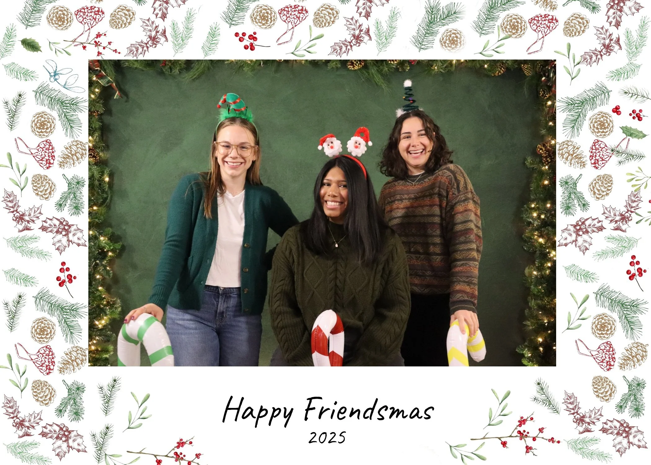 Three friends smiling in Christmas-themed headbands and sweaters, standing against a green backdrop decorated with holiday greenery and fairy lights, holding large candy canes, framed with holiday illustrations and the words "Happy Friendsmas".