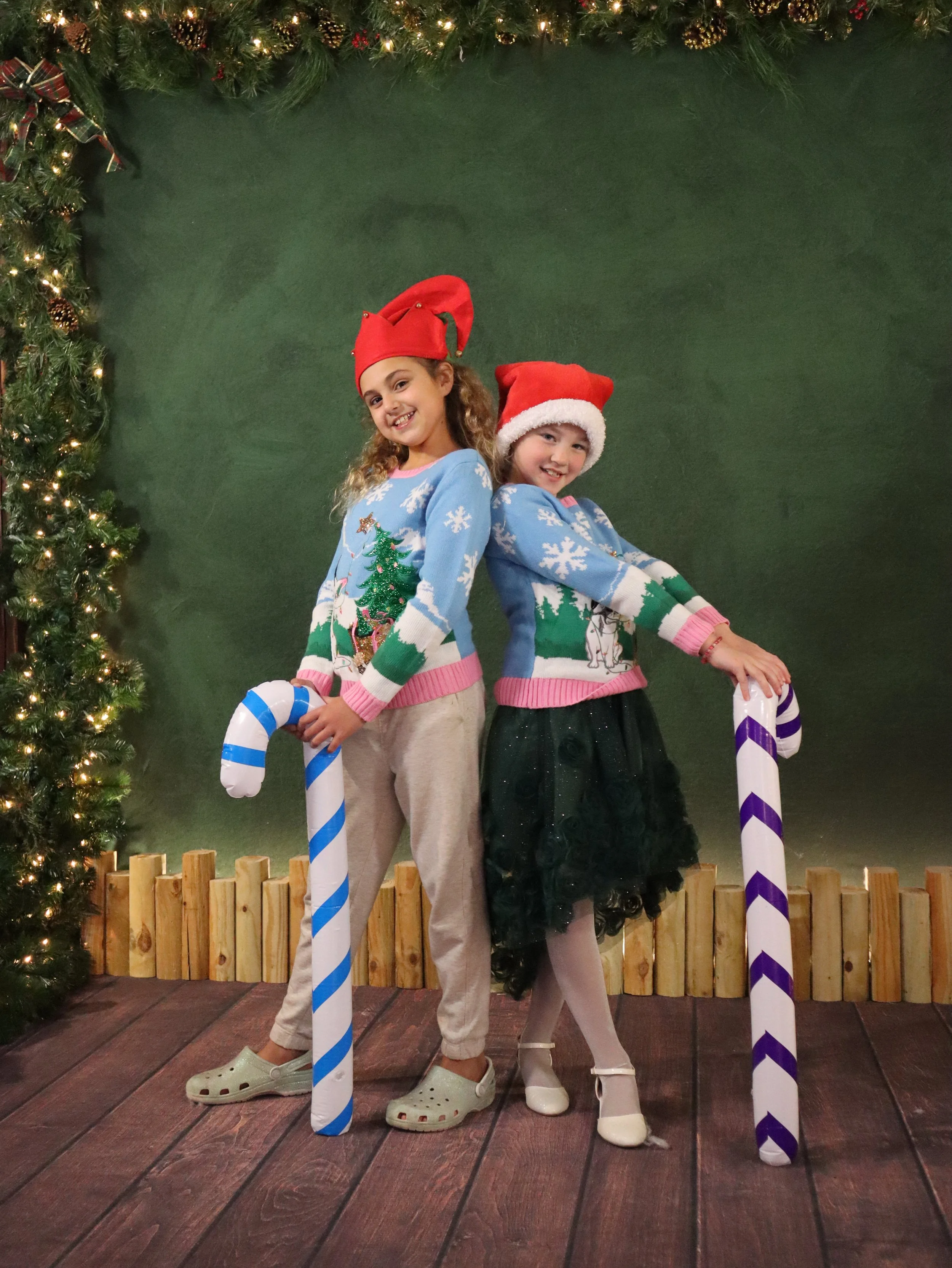Two young girls dressed in Christmas sweaters and Santa hats, holding candy cane props, standing in front of a Christmas-themed backdrop with a decorated tree and string lights.
